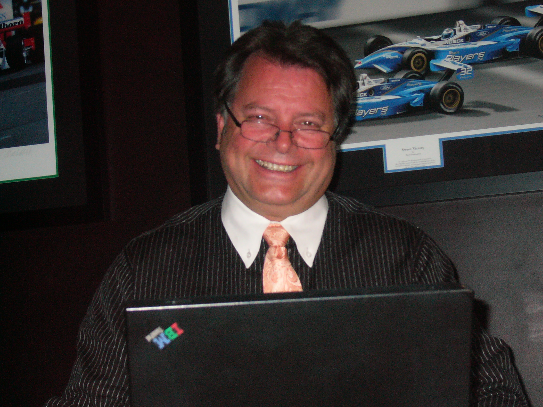 Colin in front of a black laptop. He is wearing glasses, a black striped dress shirt and a pink tie.