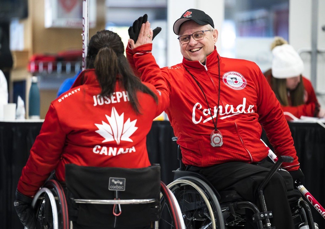 Two athletes in wheelchairs wearing Team Canada jackets exchange a celebratory high‑five in a curling event.  Two athletes in wheelchairs wearing red Team Canada uniforms compete in wheelchair curling on an indoor ice rink.