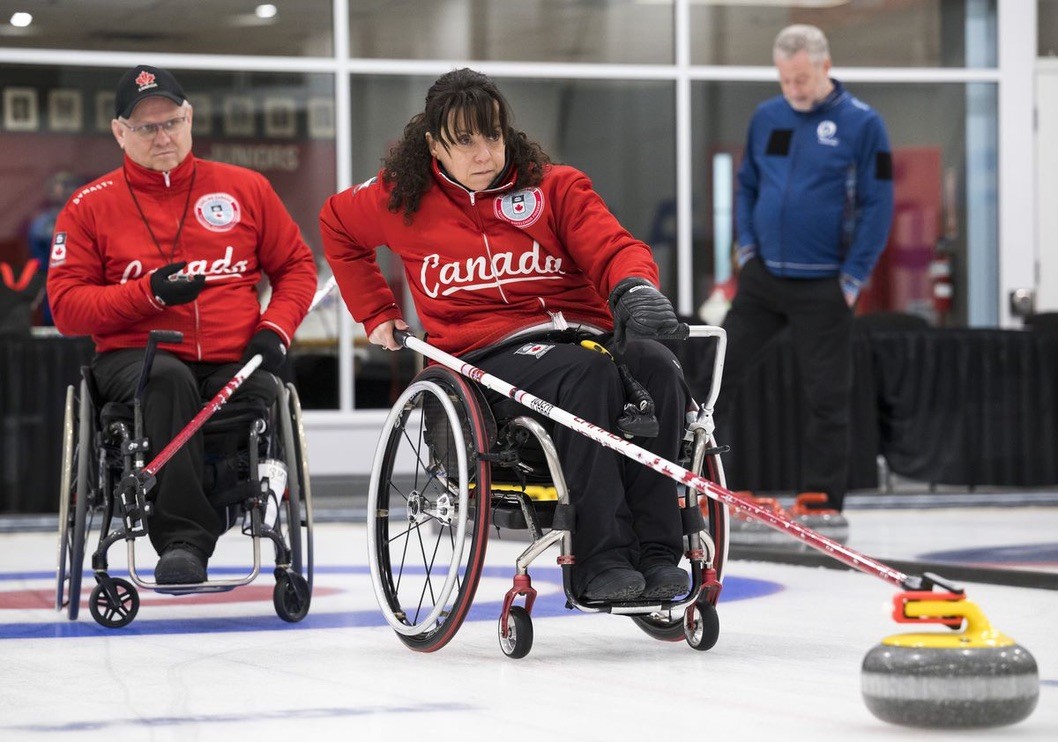 Two athletes in wheelchairs wearing red Team Canada uniforms compete in wheelchair curling on an indoor ice rink.