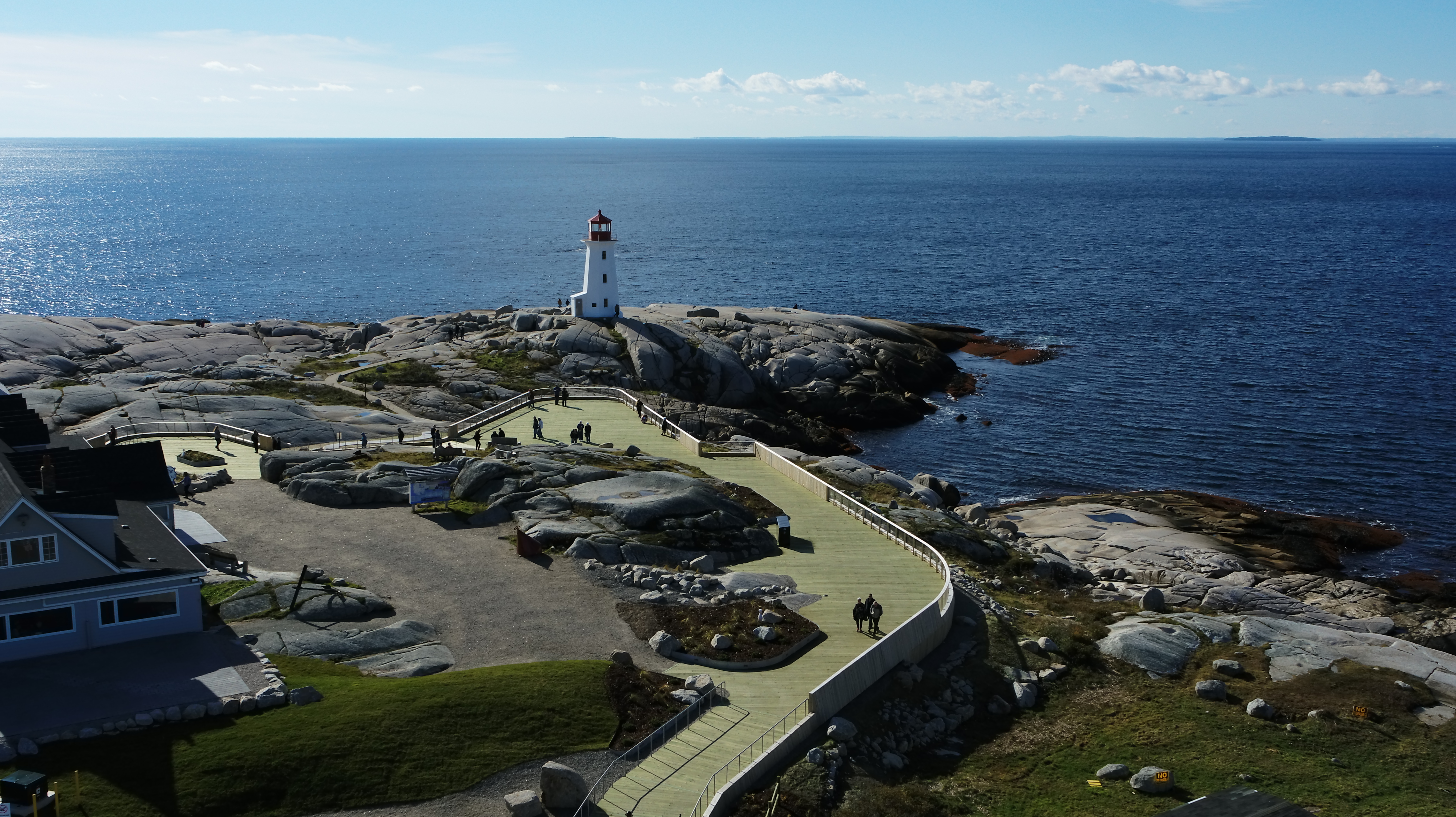 Aerial view of the Peggy's Cove viewing deck. The viewing deck is a large, wooden accessible platform that wraps around the large rocks that surround the red and white lighthouse, which overlooks the ocean. There is a building near the trai.