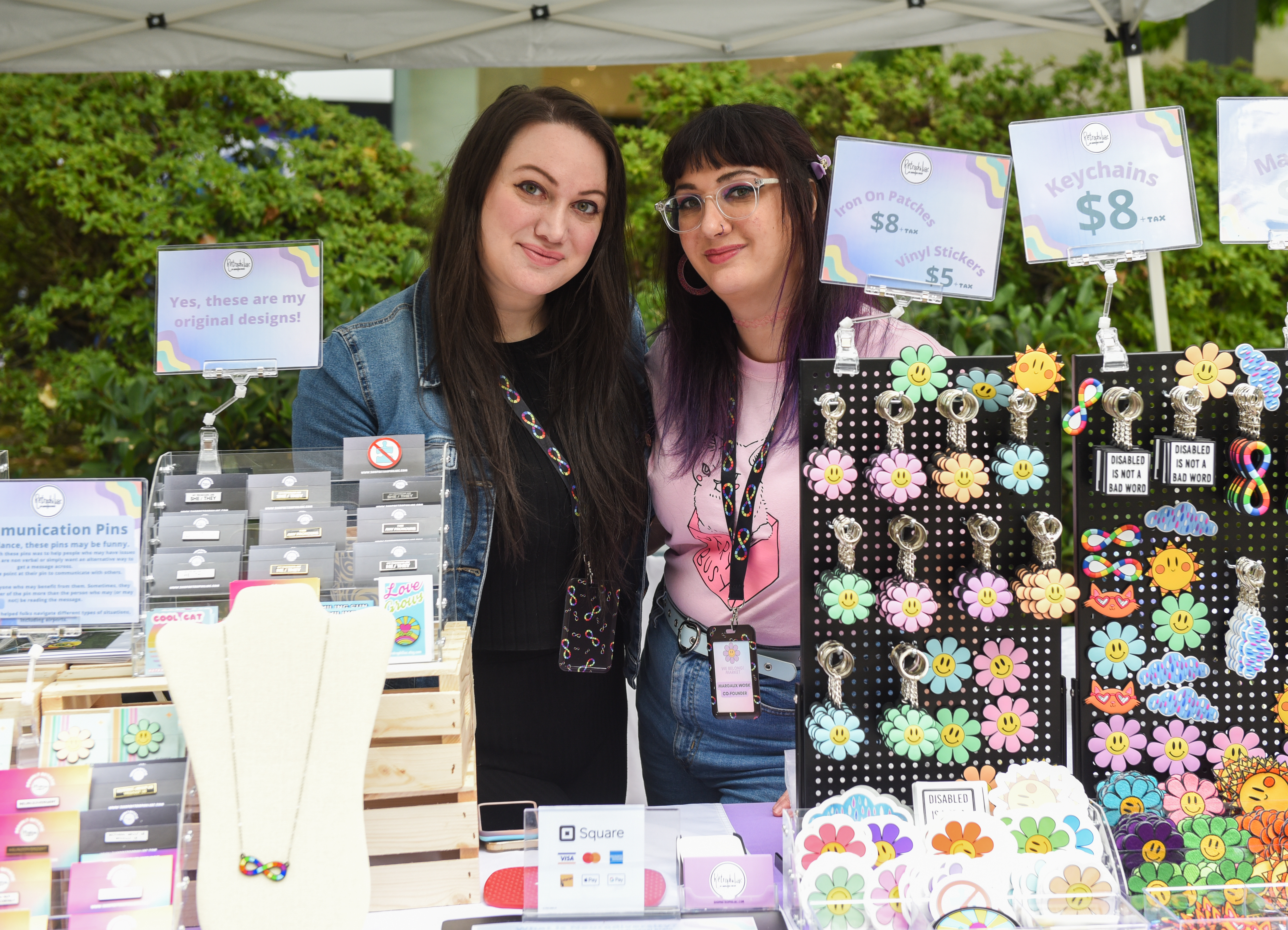 We Belong! Festival founder Margaux Wosk on the right with sister Becky Wosk. Margaux has long dark hair with purple in it, is wearing a pink t-shirt and has glasses. Becky has long dark hair and is wearing a black outfit with a denim jacket. They are in front of a stand of items that are for sale.