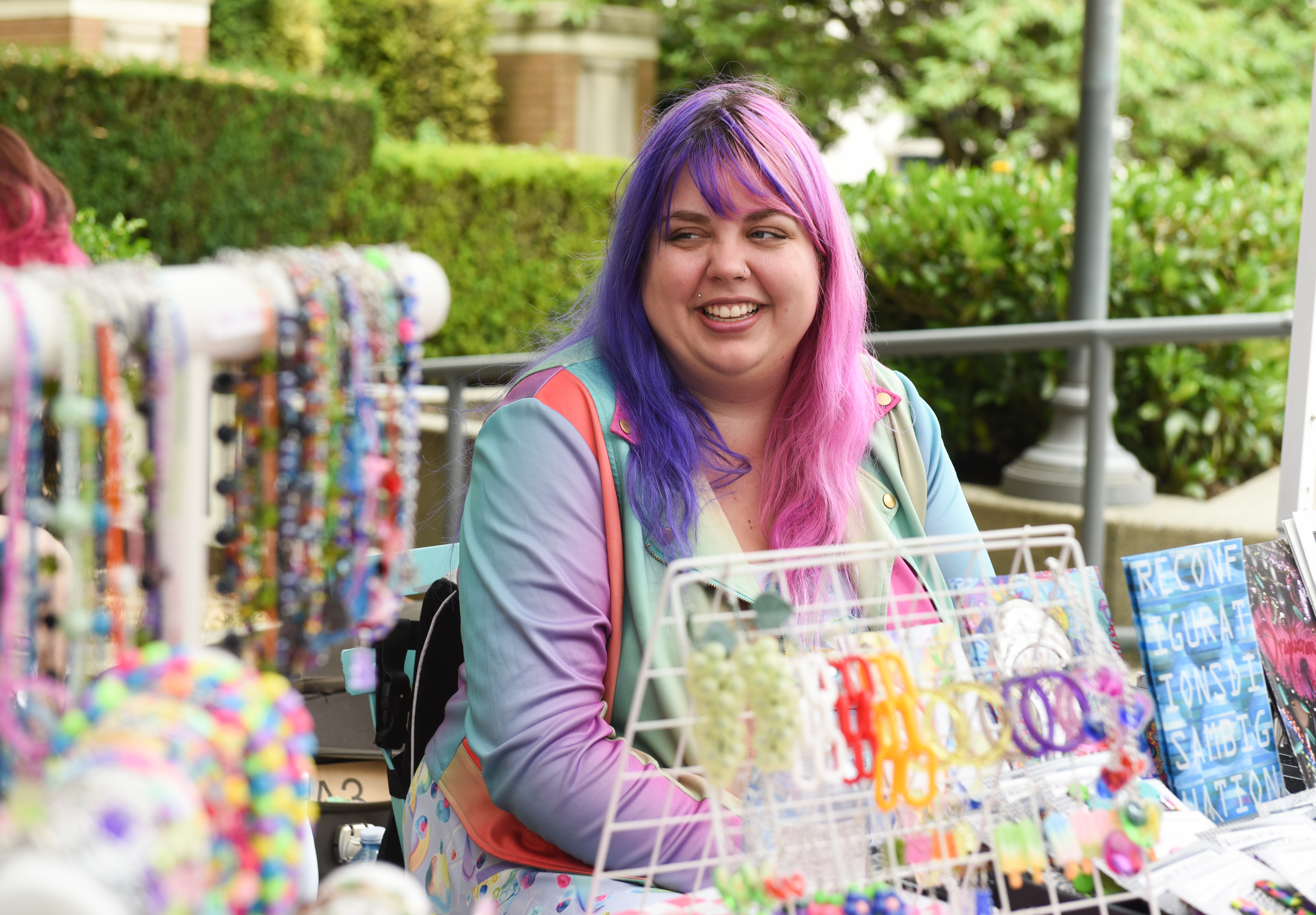 Kirsten Hatfield, who has long hair that is half purple and half pink and is sitting at a table selling jewellery.