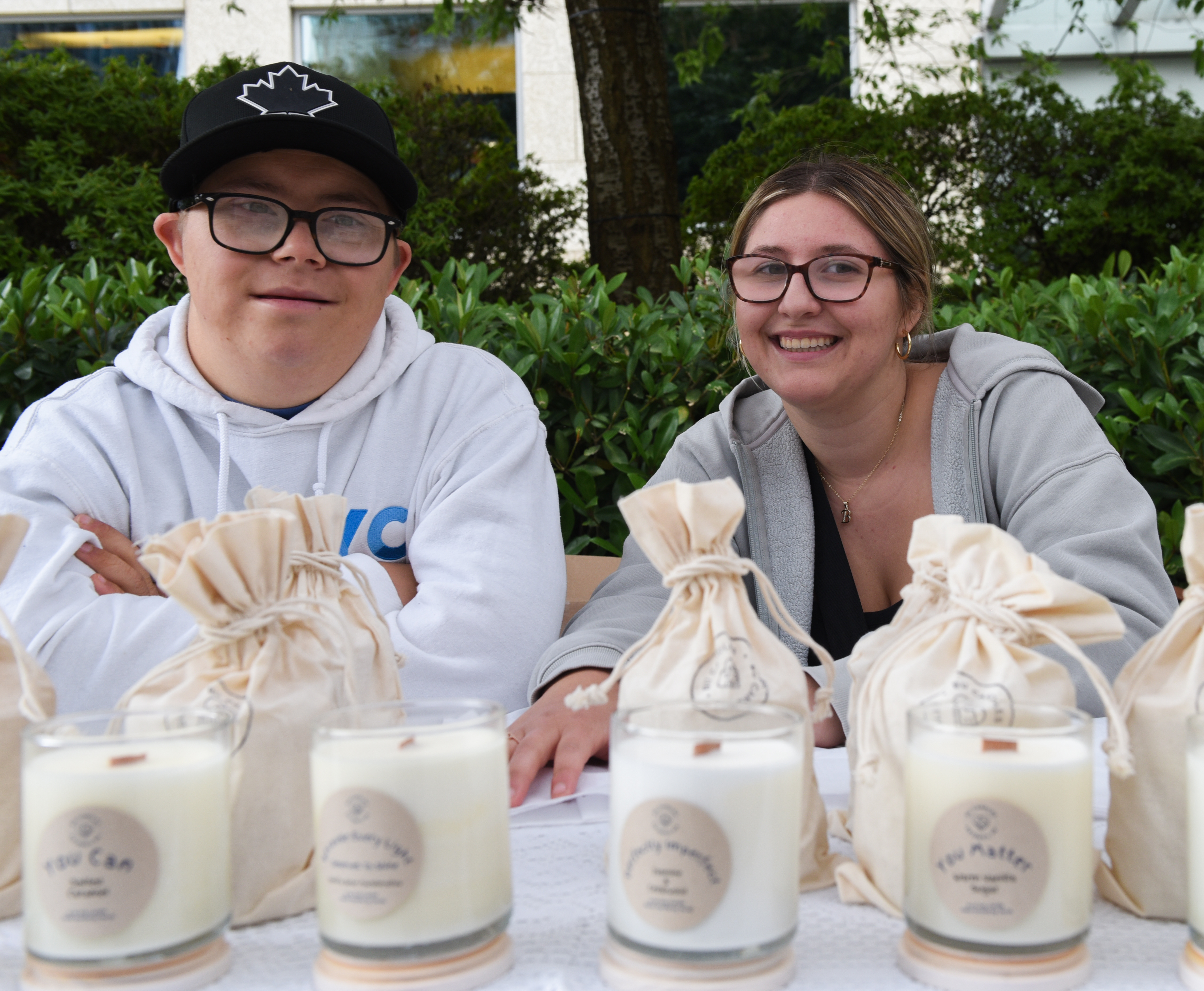 Two people sitting at a stand selling candles. The person on the left is wearing a black ball cap, a white hoodie and has glasses. The person on the right has glasses and brown and blonde hair, and is wearing a grey sweater with gold earrings and a gold necklace.