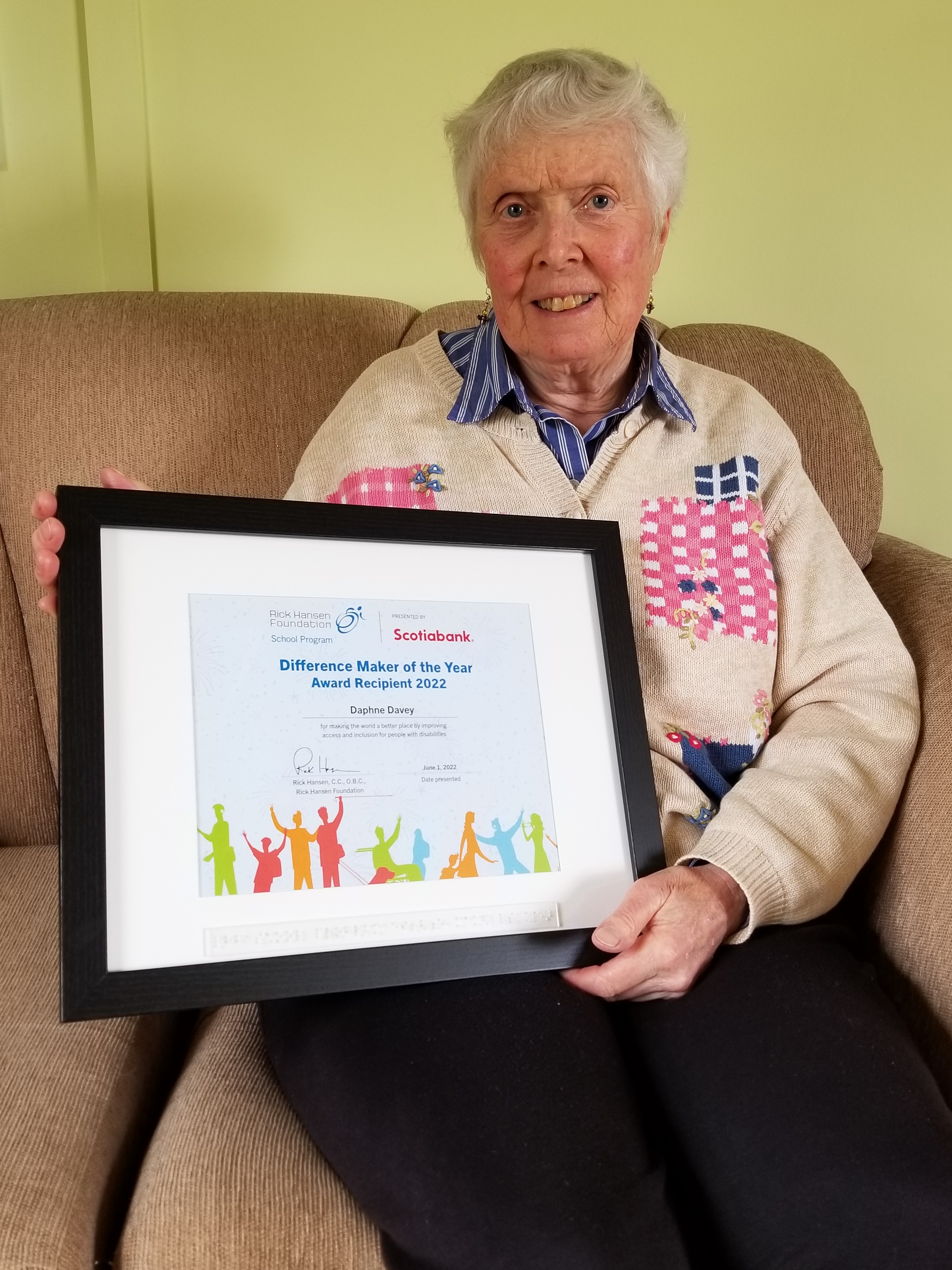 Woman sitting on a brown couch holding a Different Maker Award certificate. She is wearing a white sweater with pink and blue patches.