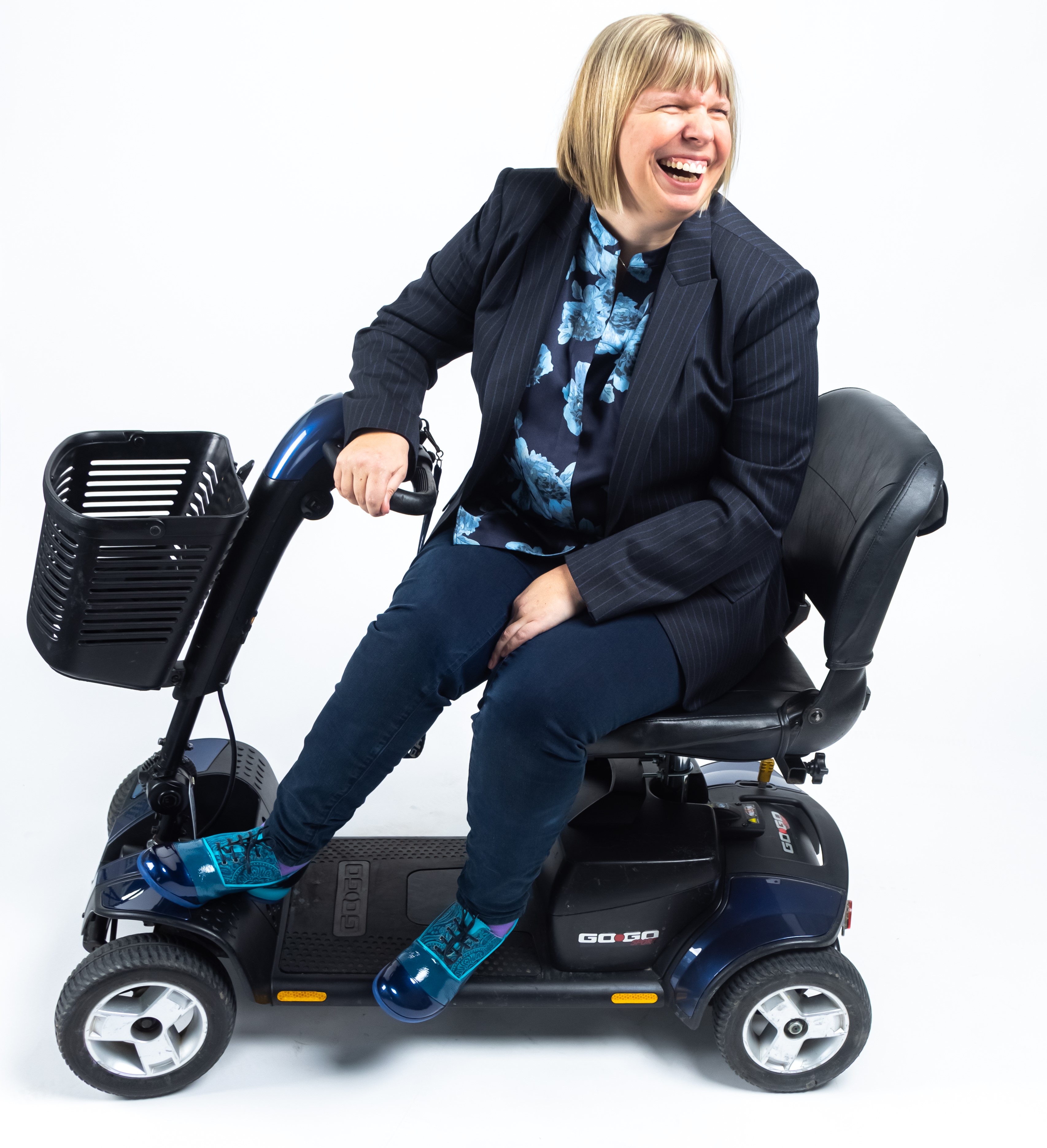 Darby Lee Young who has short blonde hair cut in a bob. Darby is wearing a striped blazer, a floral shirt and shiny blue shoes. She is laughing and is using a motorized mobility device.