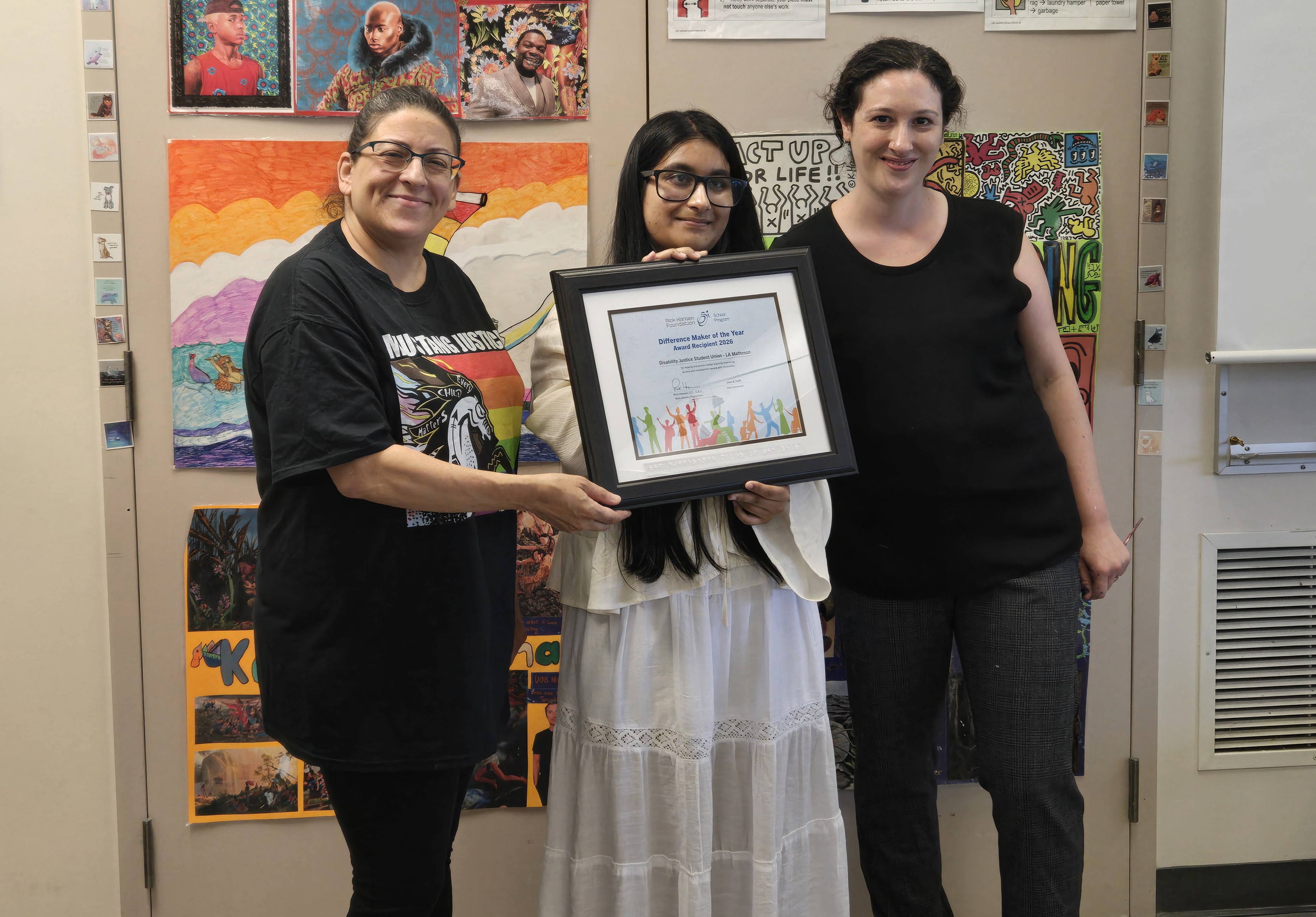 Three people standing indoors beside student artwork, holding a framed “Difference Maker of the Year Award Recipient 2026” certificate together.