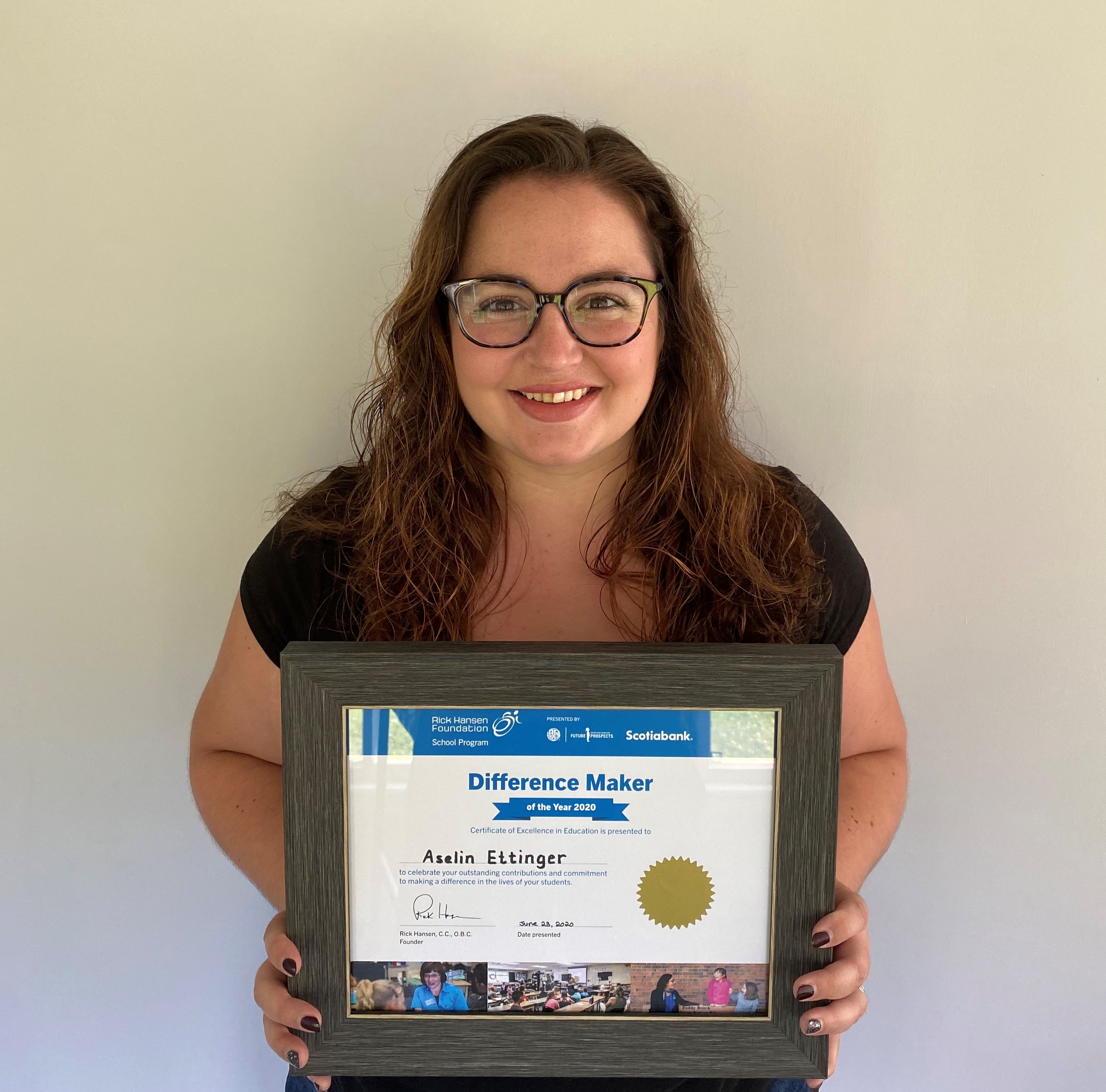 Young woman brown hair wearing glasses smiles and holds her certificate.