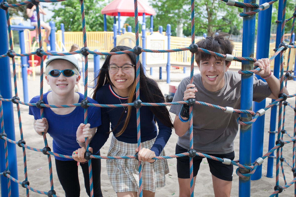 Elena at the park with her brother and sister. The three are looking through a climbing net and smiling 