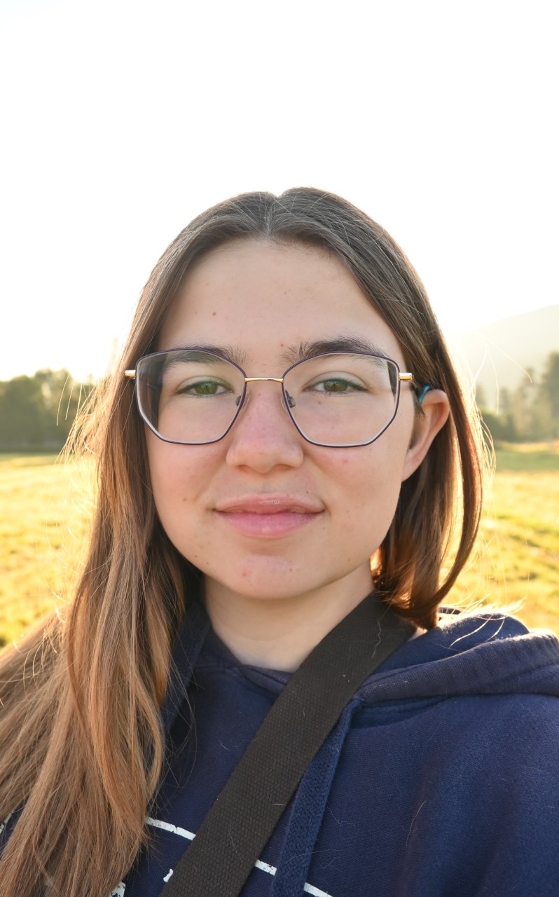 Headshot of Elise Doucet, she is wearing octagonal framed glasses and a blue hoodie, and has shoulder length brunette hair. The sun is shining behind her in a rural setting.