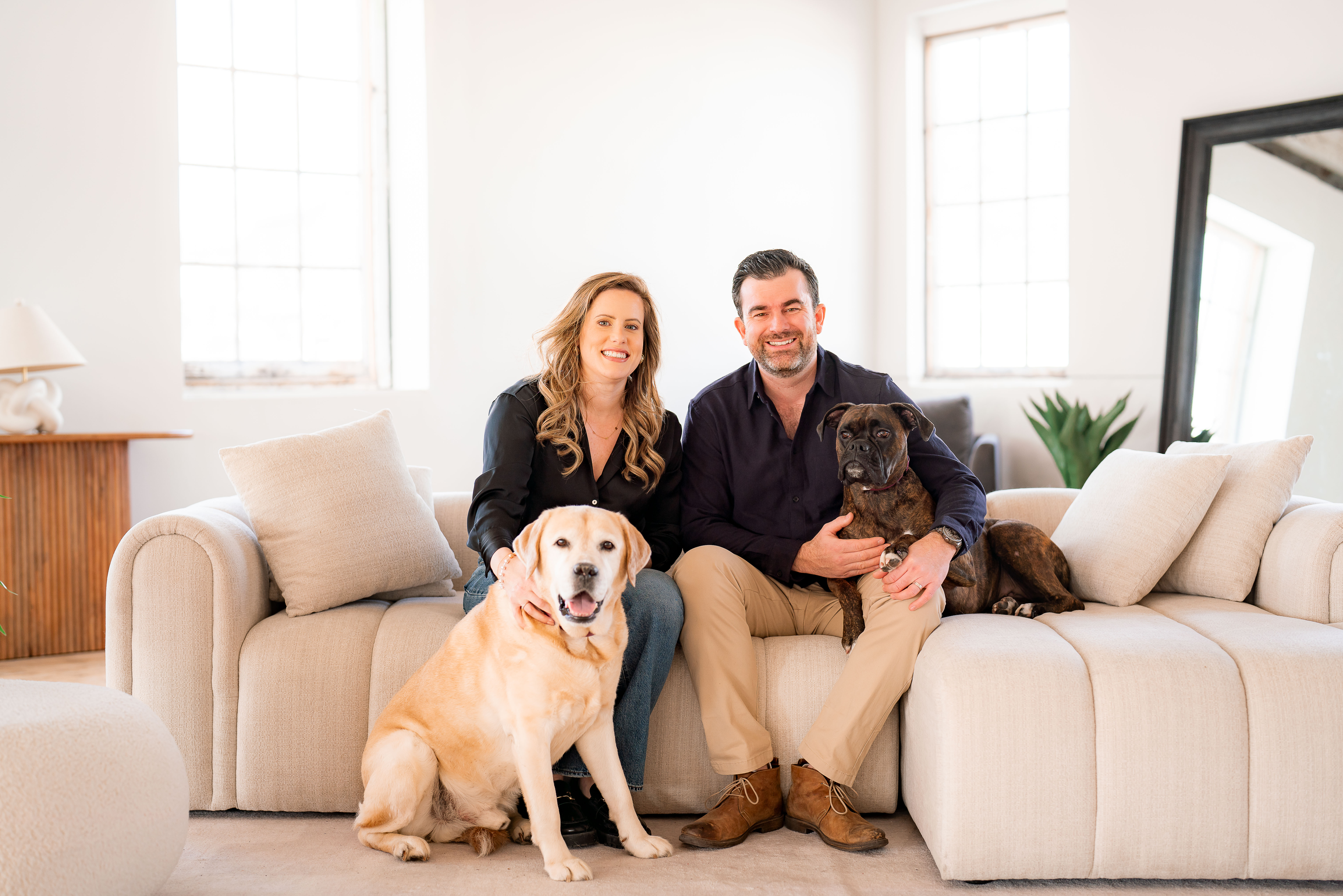 A man and a woman dressed in formal attire sit on a large white couch, hugging a large dog and holding a smaller one