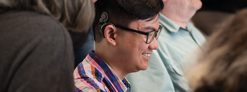 A person with a cochlear implant smiling. He has black hair and glasses.