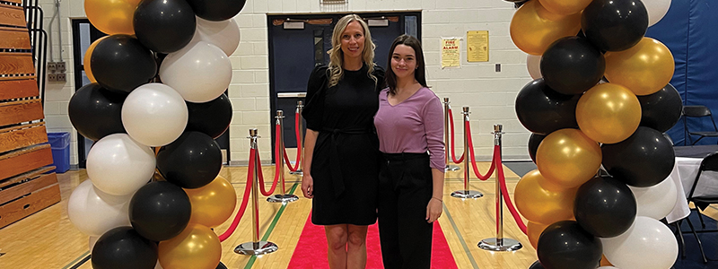 A 2023 Difference Maker standing under a balloon arch with her teacher at a high school prom for students with disabilities.
