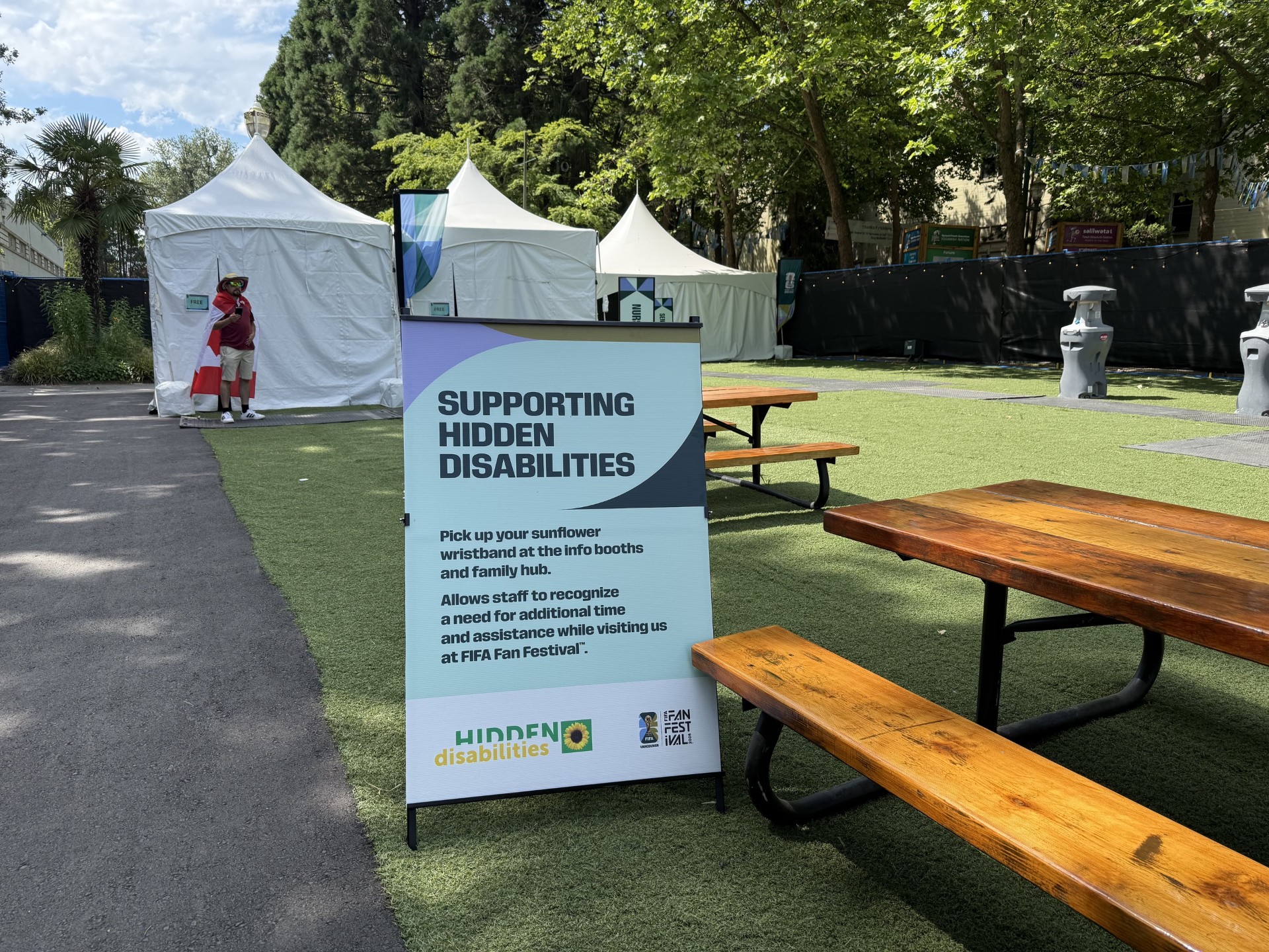 Sign reading “Supporting Hidden Disabilities” beside picnic tables and festival tents, explaining the availability of sunflower wristbands for visitors who may need additional assistance.