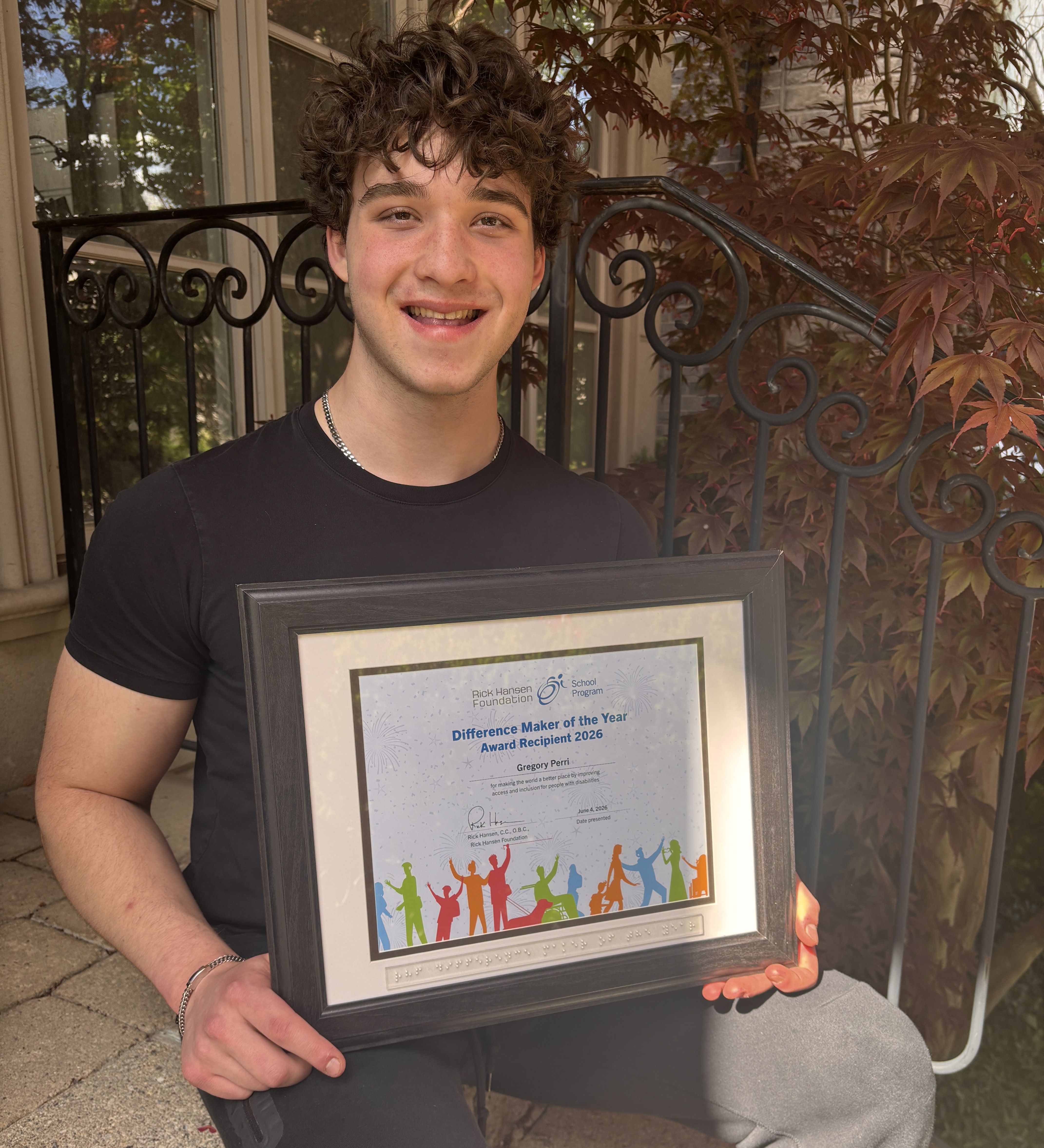 Young man with short curly hair standing outdoors near a metal railing and plants, holding a framed “Difference Maker of the Year Award Recipient 2026” certificate.