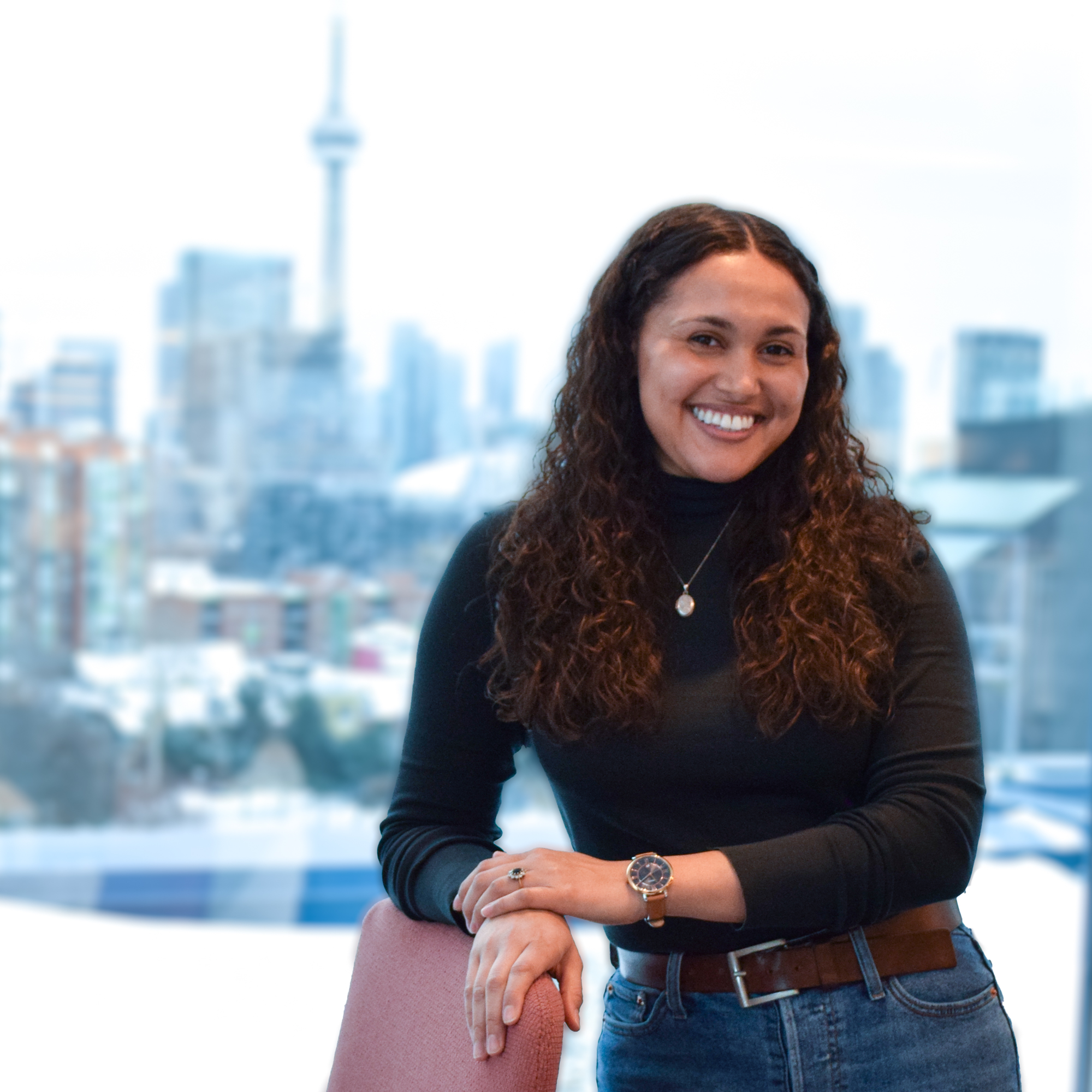 Haley Rae Dinnall-Atkinson, who is a woman with long brown curly hair smiling and leaning on a brown chair. She is wearing a black turtle neck sweater and blue jeans. Behind her is an open window to a city landscape.