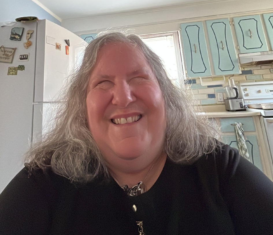 A woman who is blind smiles broadly, wearing a black t-shirt and with short hair. Behind her, a kitchen is visible, featuring cabinets and a fridge.