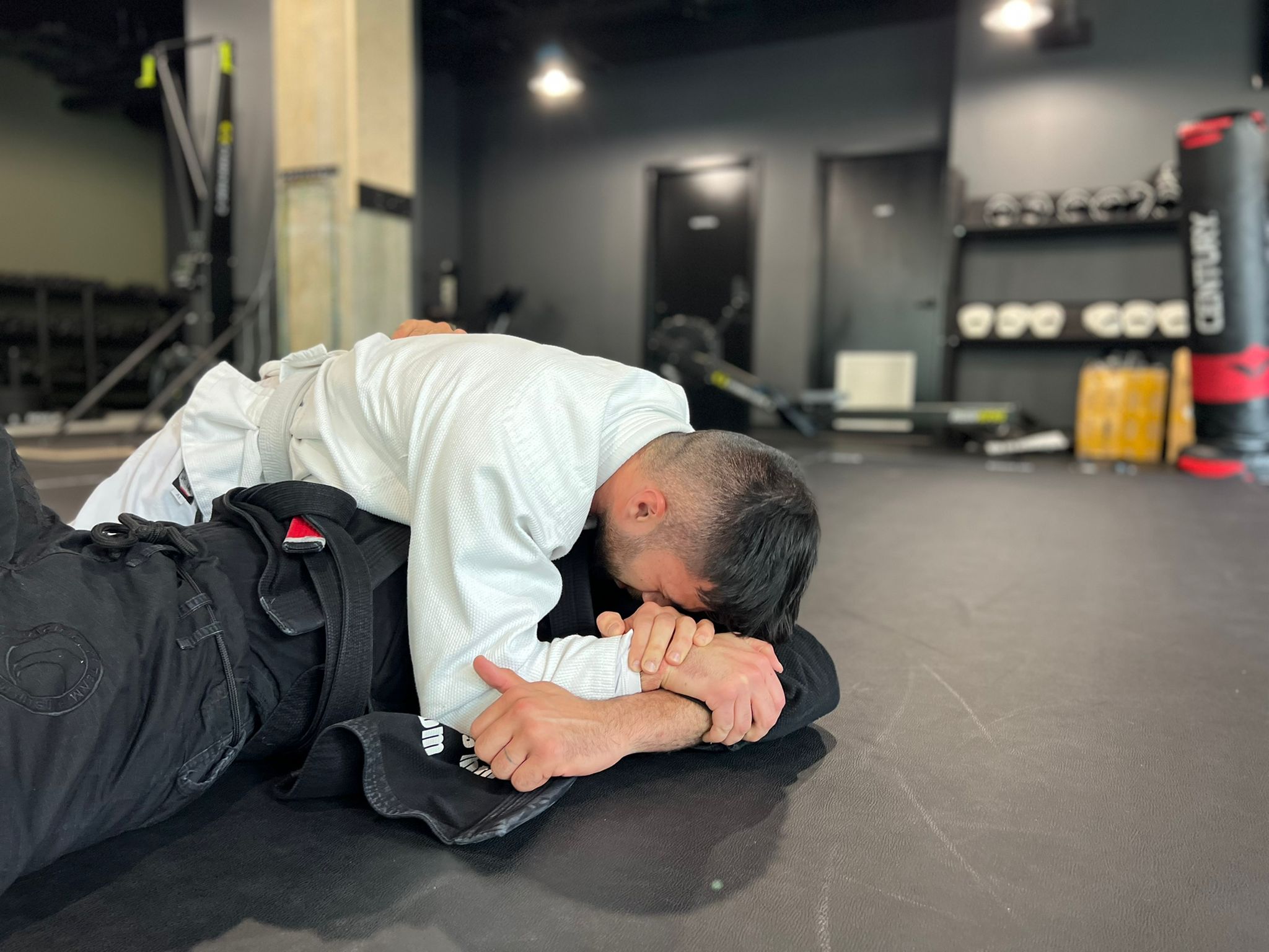 Two people participating in a martial arts match. A person wearing a white uniform is pinning down a person in a black uniform.