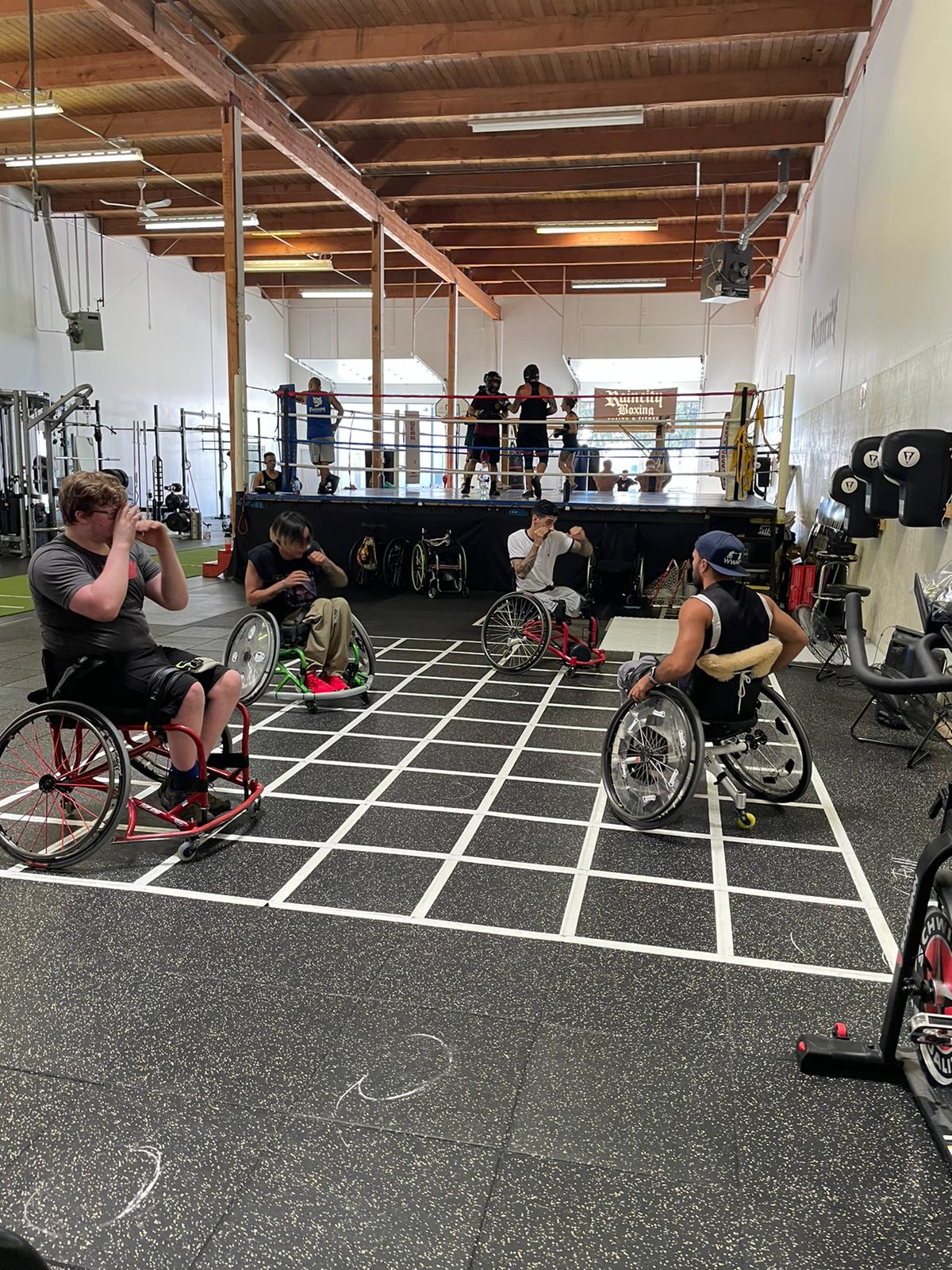 A boxing gym filled with people who are using wheelchairs training inside a large square grid made out of tape on the ground. 