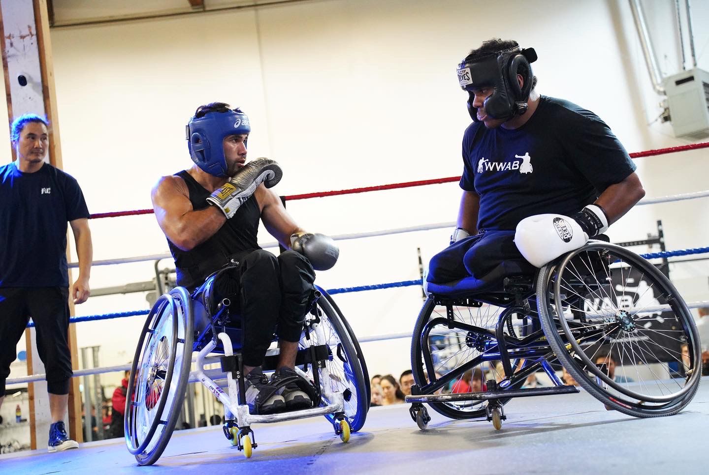 Two people who are using wheelchairs boxing in a ring. One person is wearing a blue helmet and one person is wearing a black helmet.