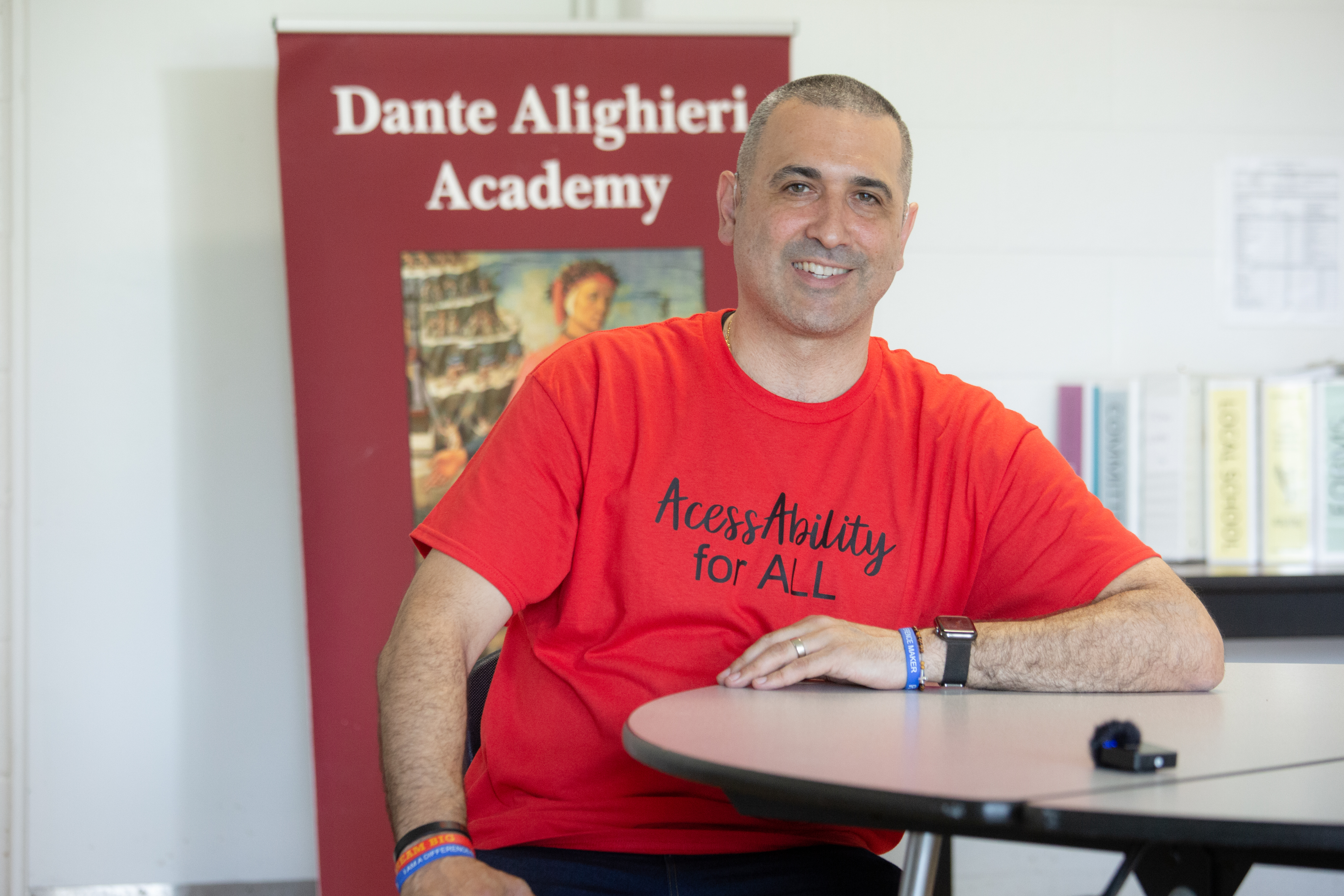 Angelo sitting at a table smiling and wearing a red t-shirt that says AccessAbility for all. There is a banner behind him that says Dante Aligheri Academy