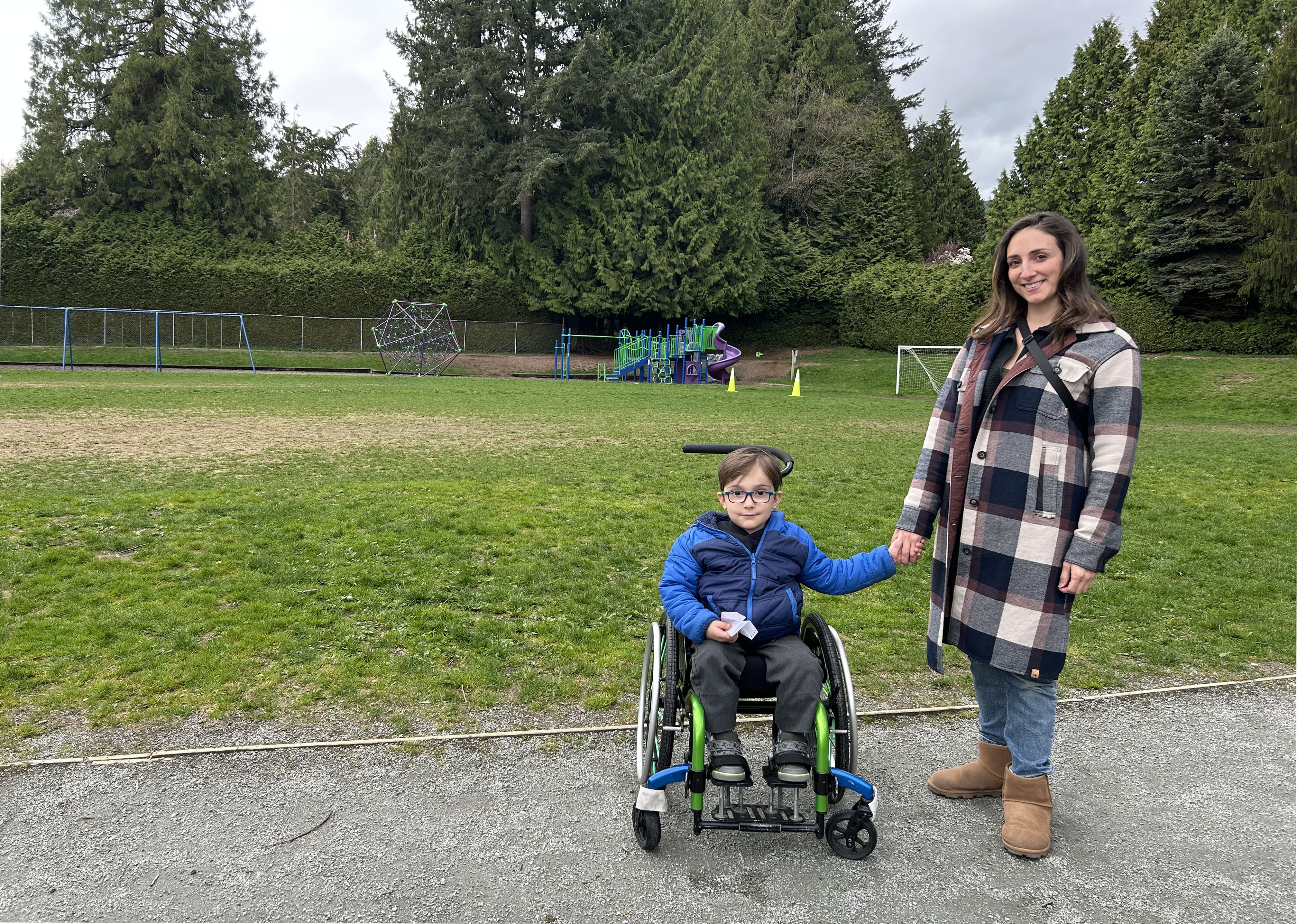 A woman and a child in a wheelchair pose in front of a playground. They are smiling and holding hands
