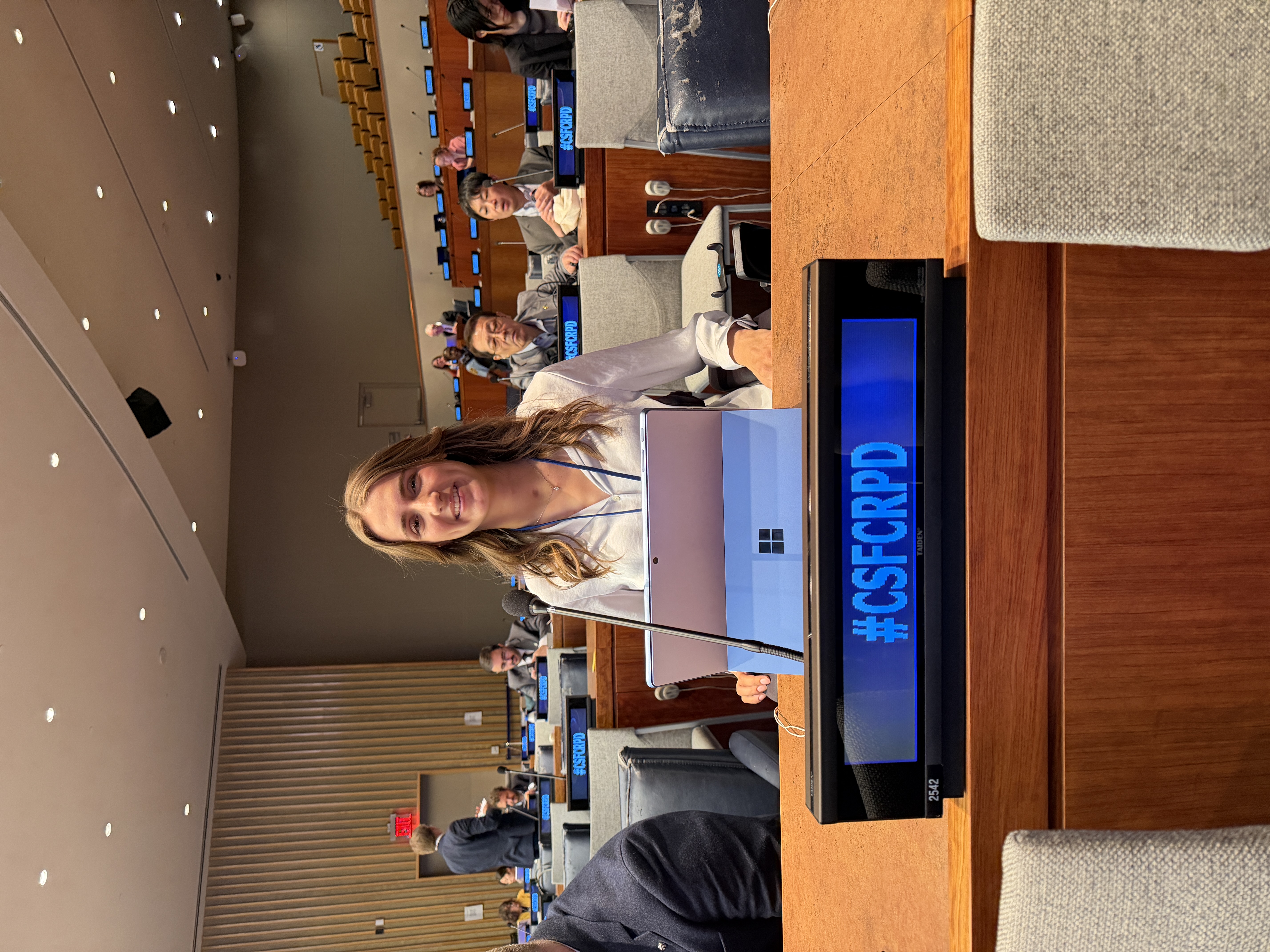 Young woman with long wavy brown hair seated at a conference desk in a large assembly hall, using a laptop. A microphone and a display panel reading “#CSFCRPD” are positioned in front of her, with delegates seated in the background.