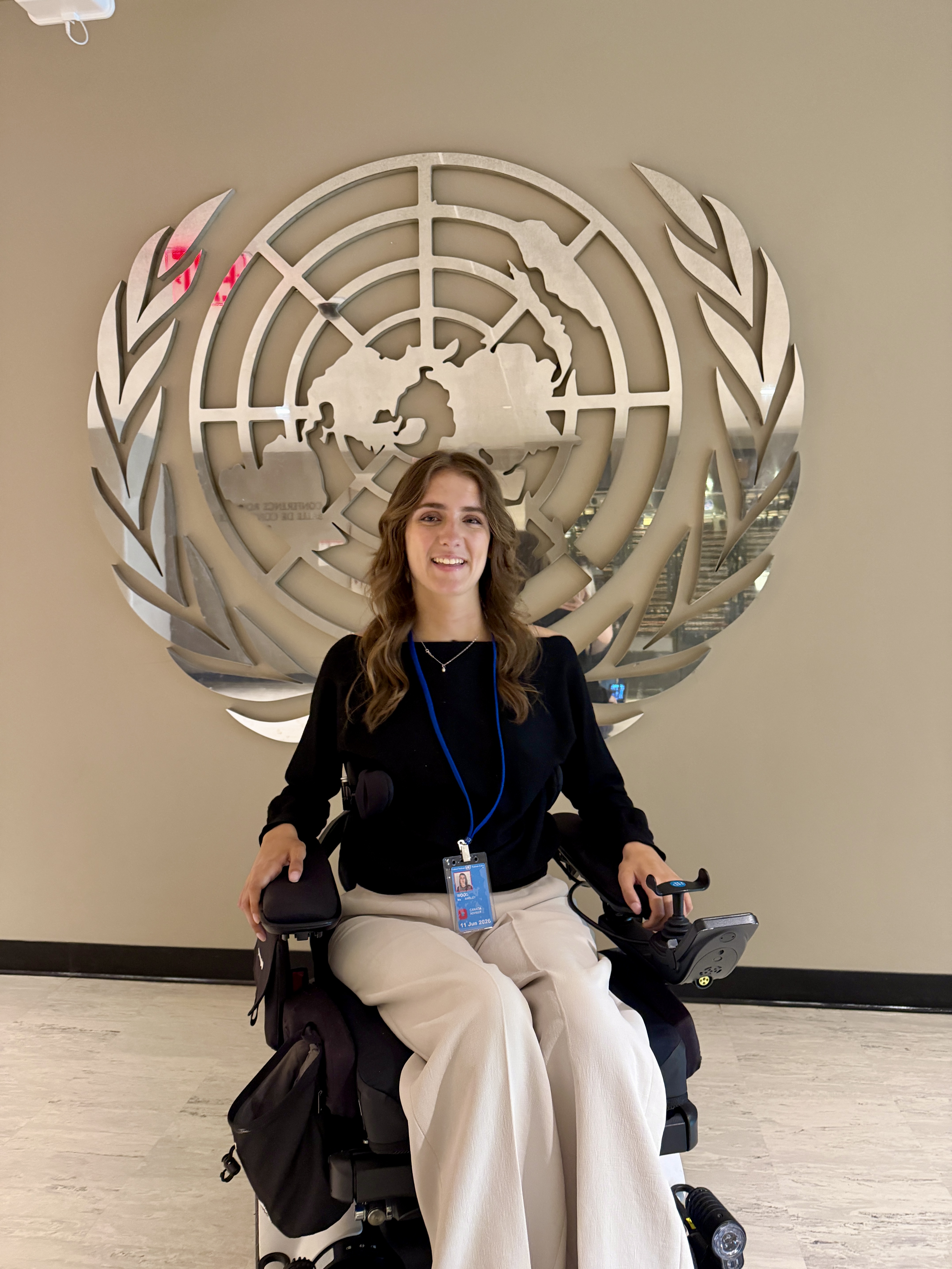 Young woman with long wavy brown hair seated in a powered wheelchair in front of a large United Nations emblem mounted on a wall. She is wearing a black long-sleeve top, light-colored pants, and a conference credential.