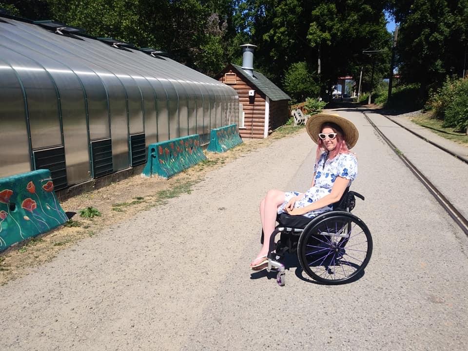 Woman using a wheelchair outdoors on a sunny pathway beside a greenhouse, wearing sunglasses, a wide-brimmed hat, and a floral dress.