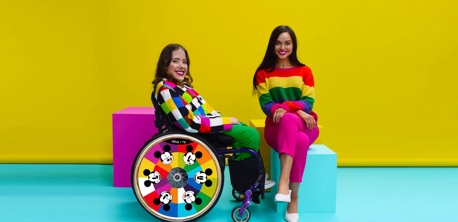 Two women, one in a wheelchair, models their disability-inclusive clothing and decorated wheels. The wheelchair model is wearing multi-coloured blazer and green pants, and her wheel has different Mickey Mouse facial expressions. The other woman is wearing a striped multicoloured sweater,  pink pants, and white shoes.
