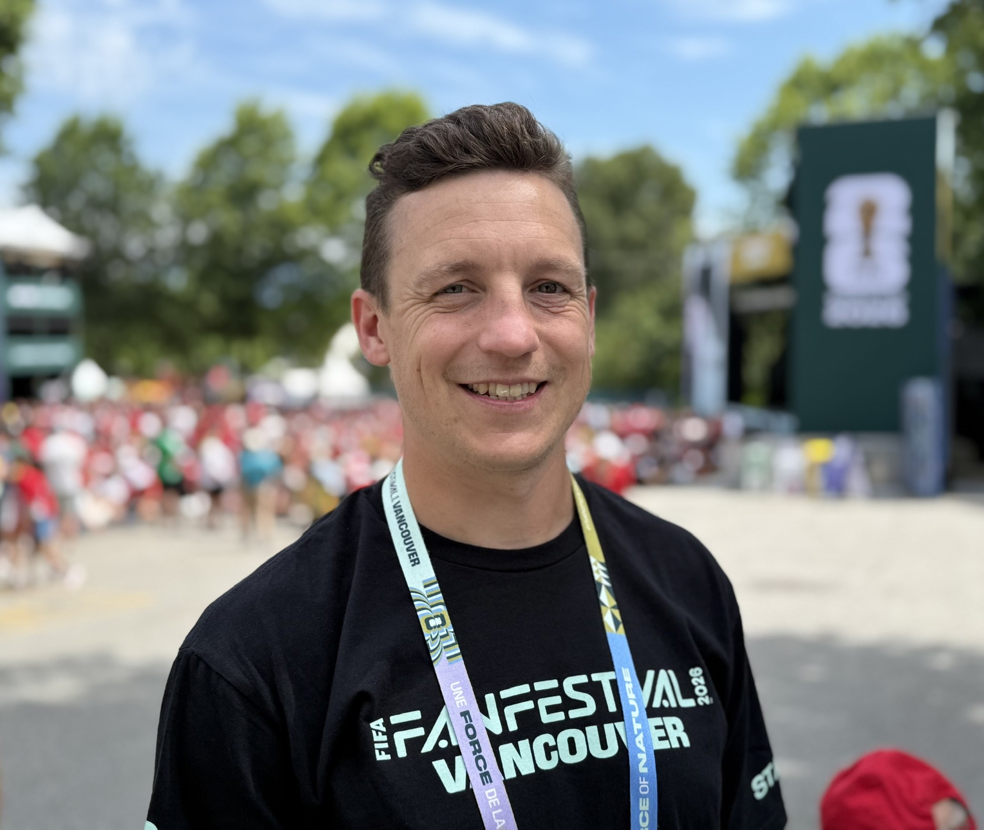 Person with short brown hair wearing a FIFA Fan Festival Vancouver staff shirt and lanyard, standing outdoors at the festival site with the crowd visible in the background.