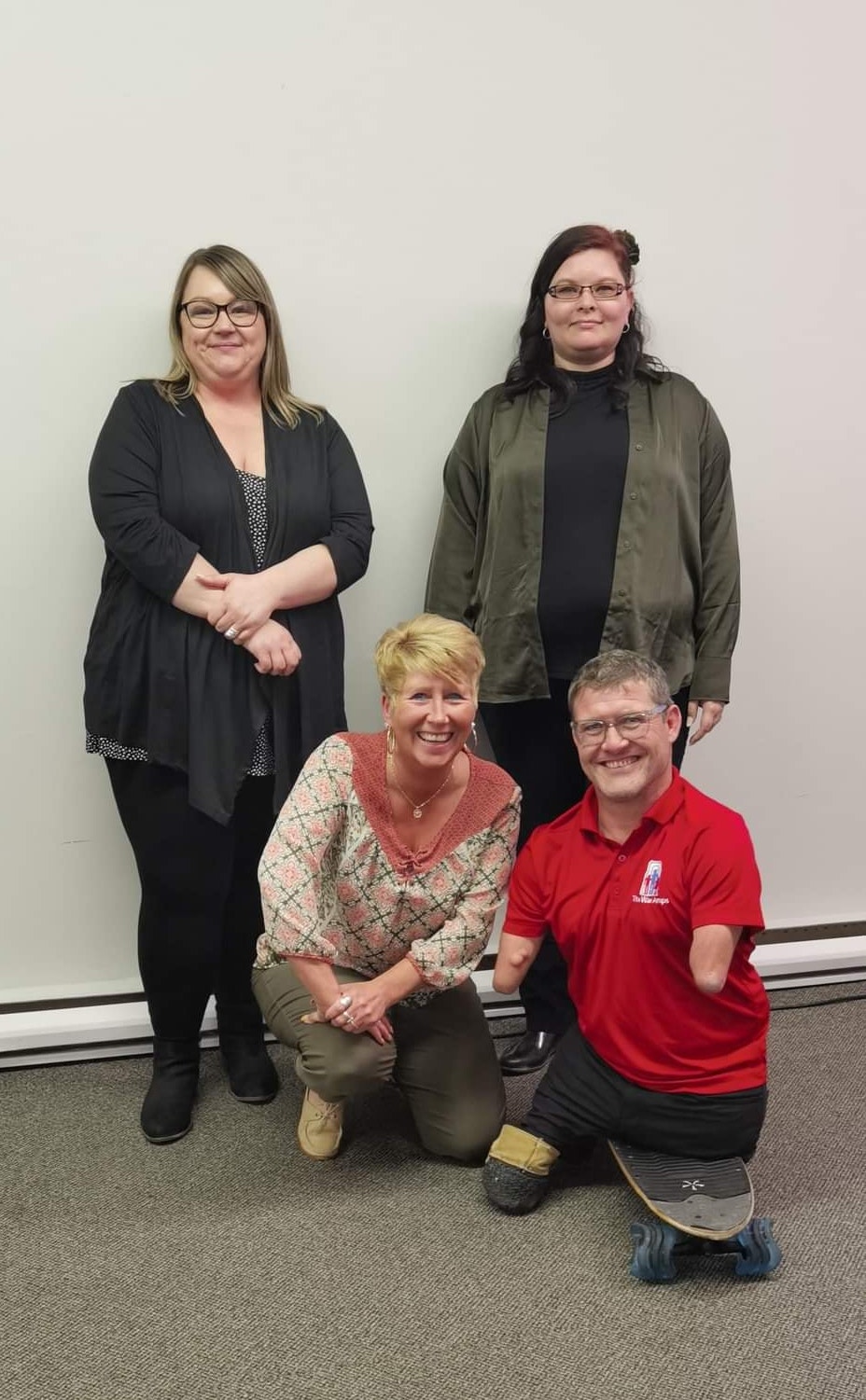 Two women standing behind another woman with short blonde hair who is kneeling beside a man who has quadruple amputation. 