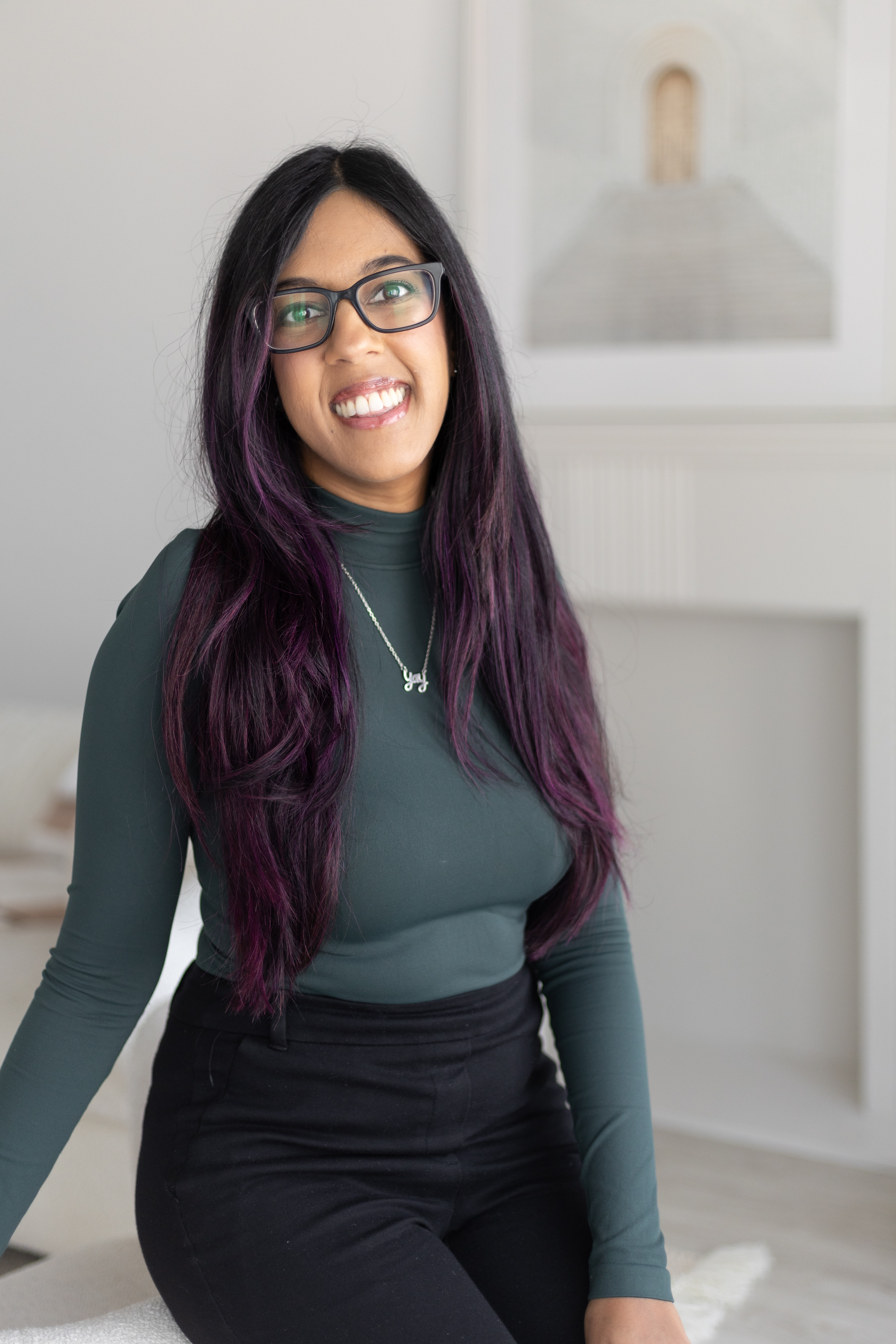 Woman with long black hair wearing glasses stands indoors near a railing, gesturing with both hands, with decorative lights and red drapery in the background.
