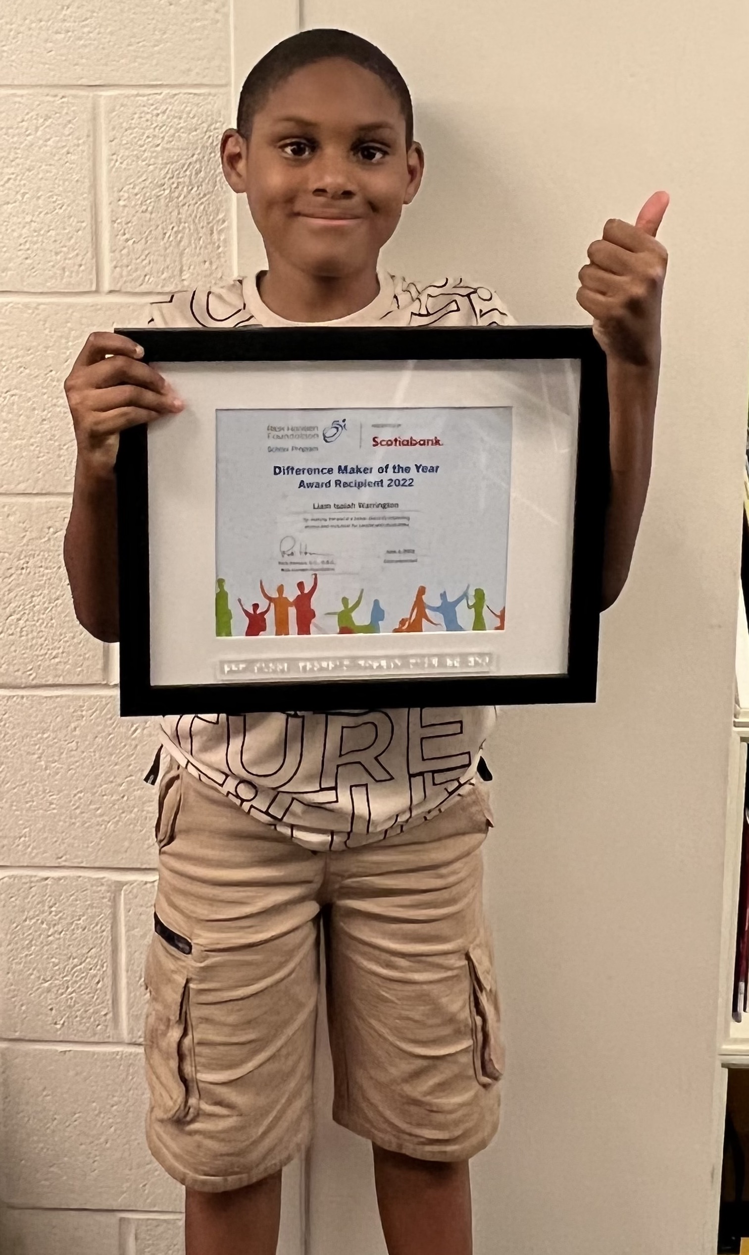 Young boy holding up a framed Difference Maker award certificate. He is wearing beige shorts and a white T-shirt.
