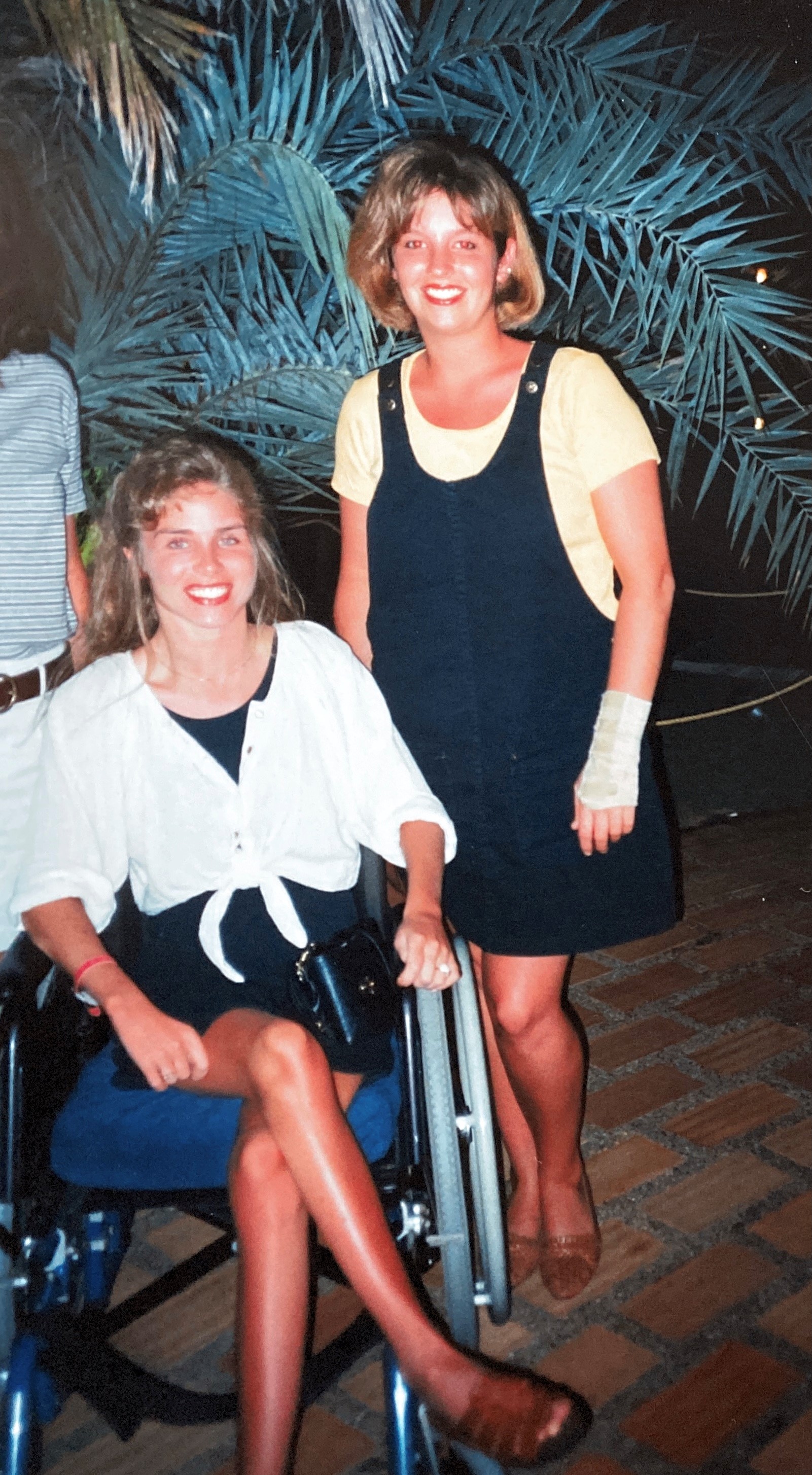 Two teenage girls, charasmaticly smiling, one using a wheelchair, in black dresses with matching brown shoes