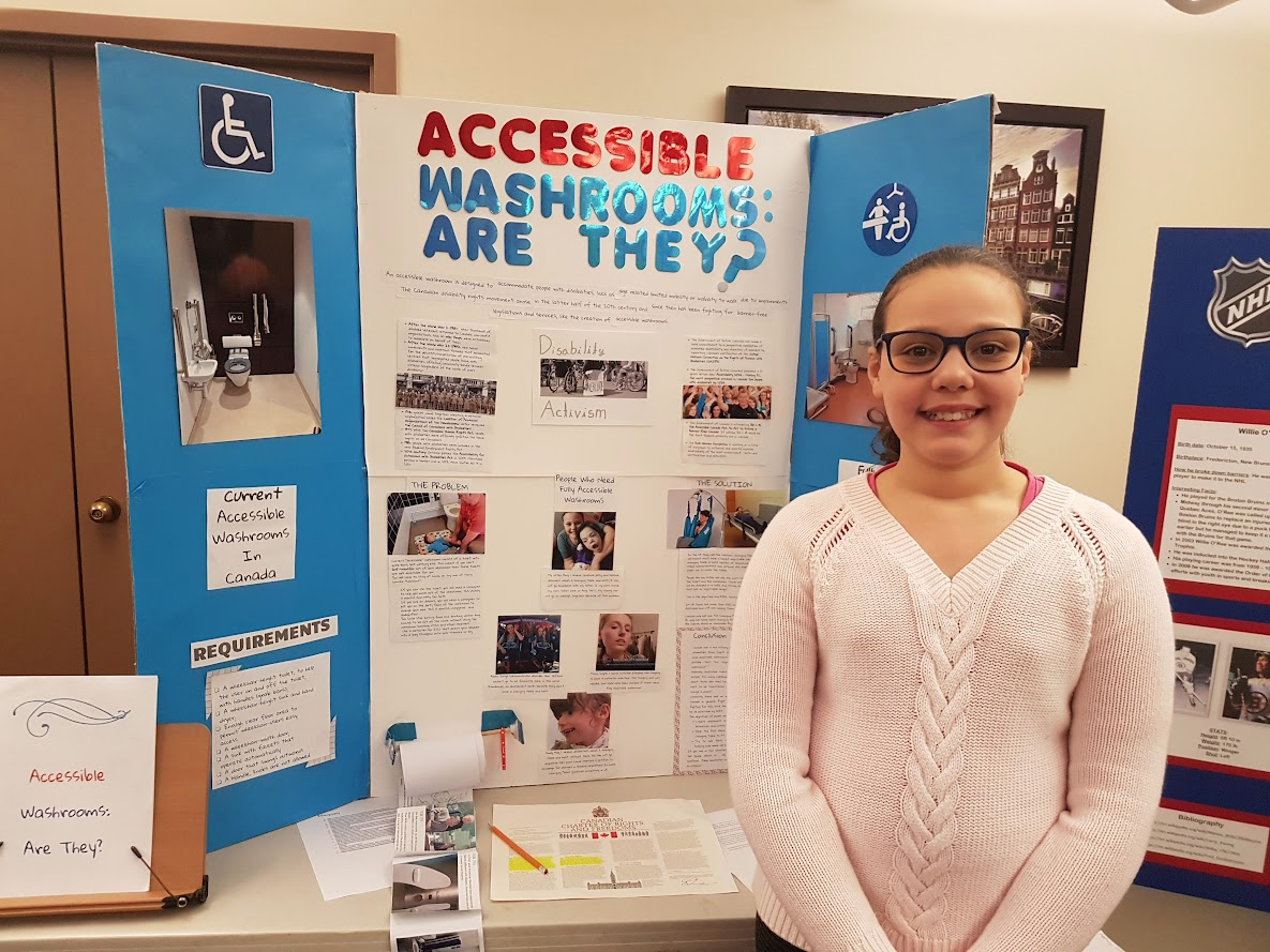Lucy Diaz, who is wearing a pink sweater and black glasses in front of her Heritage Fair project. Her project reads "Accessible Washrooms: Are They?"