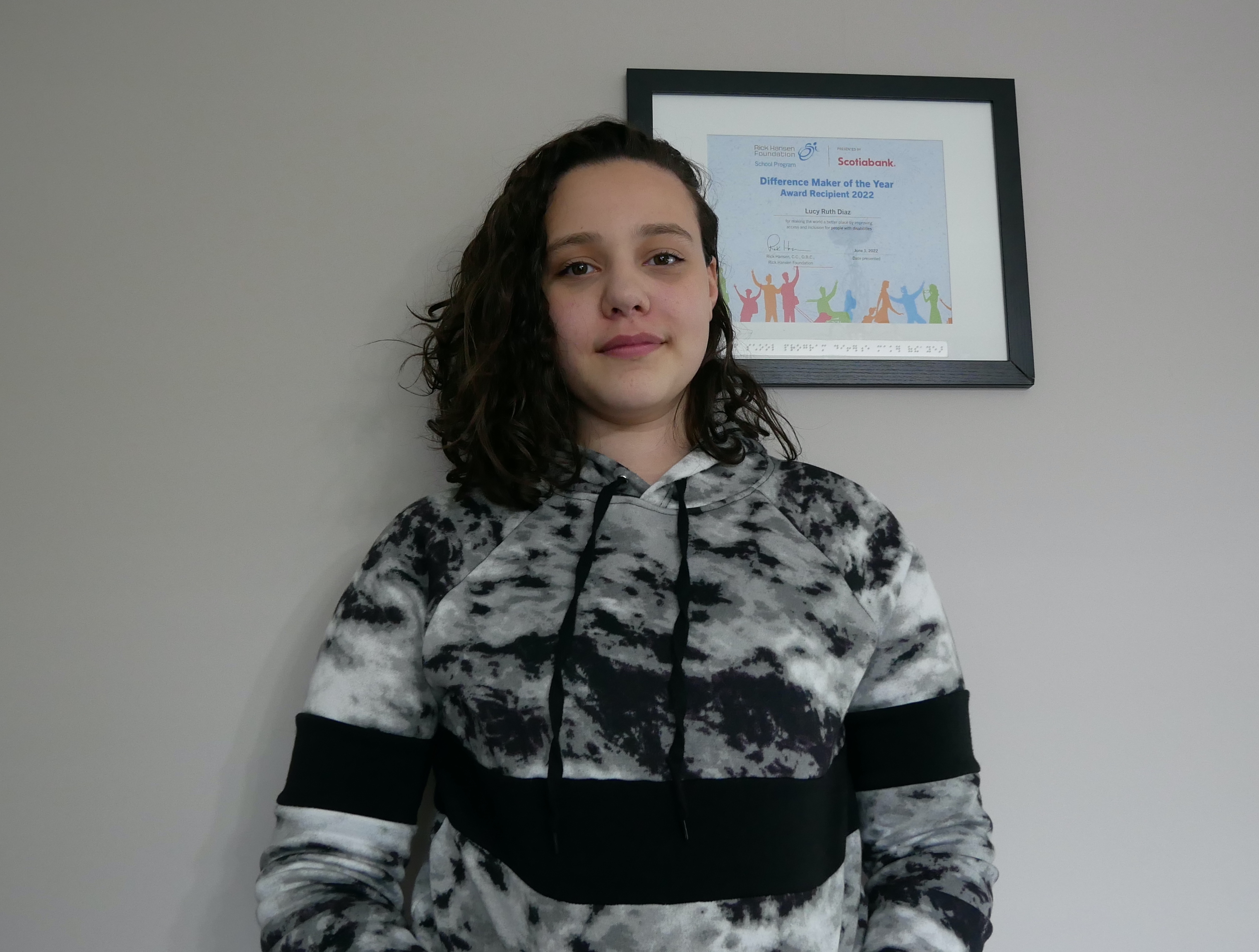 Young girl with light skin and shoulder length black curly hair. She is beside a framed Difference Maker certificate that is hanging on the wall. She is wearing a black and white tie-dye hoodie.