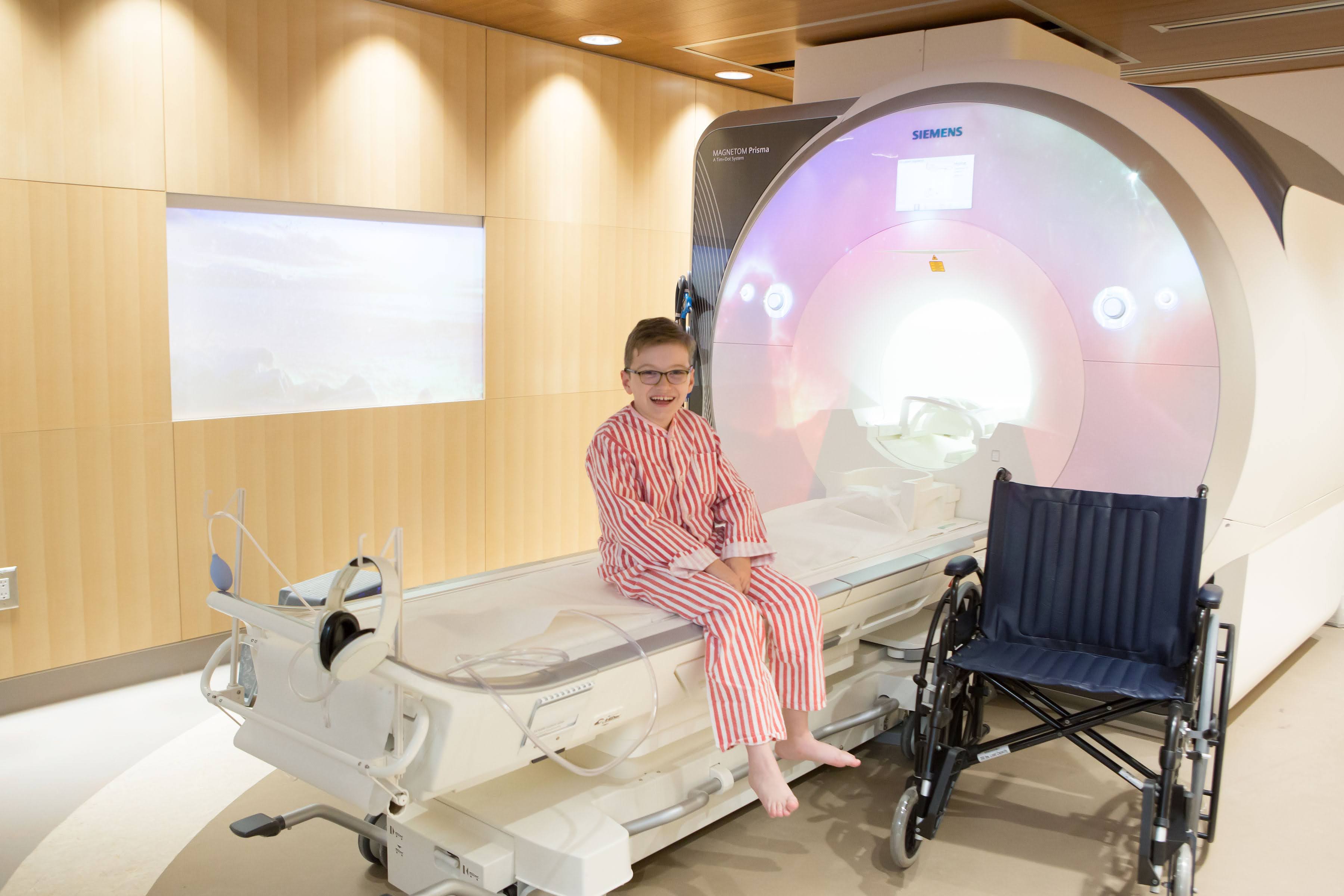 Young white boy in red and white striped pyjamas sitting in an accessible MRI suite. There is a wheelchair beside him.