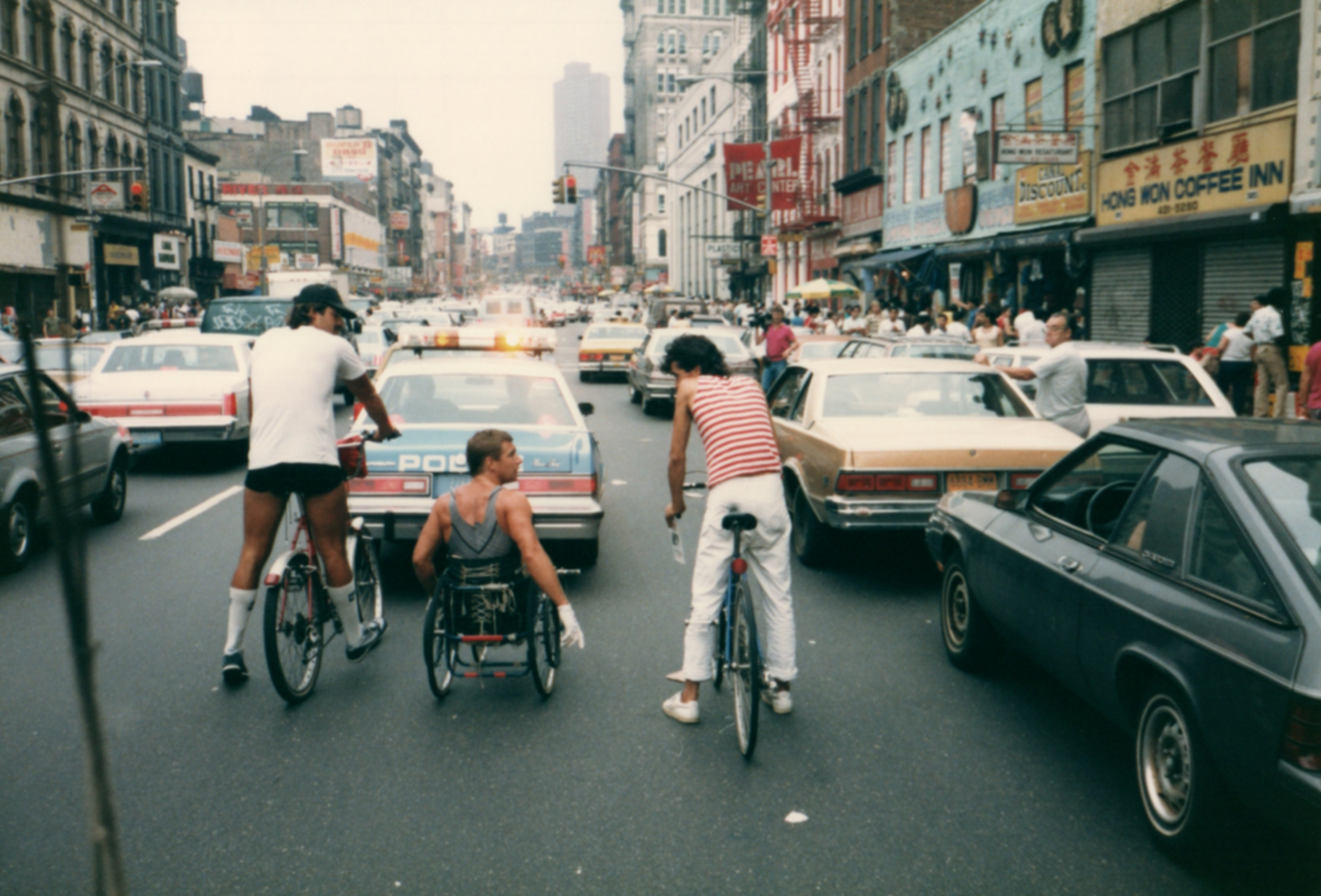 Historic street scene with a person using a wheelchair traveling alongside two cyclists in heavy city traffic, surrounded by cars and storefronts.