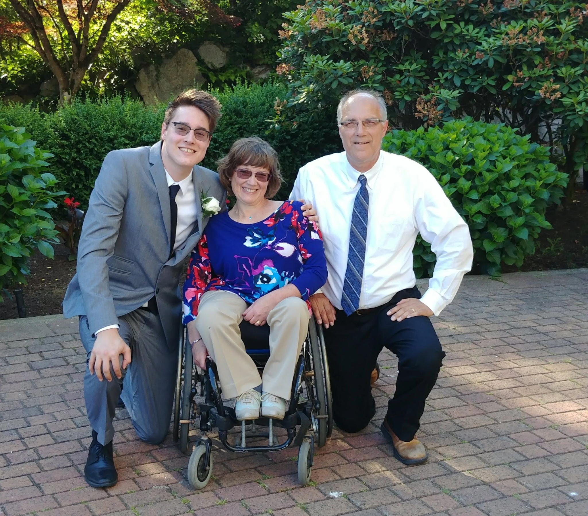 Two man, one younger than the other, kneel beside a woman seated in a wheelchair, all dressed in formal attire.
