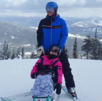 Young white girl using a bi ski who is in a pink jacket. An adult white man is standing behind her wearing skis and a blue jacket. They are on top of a snowy mountain.