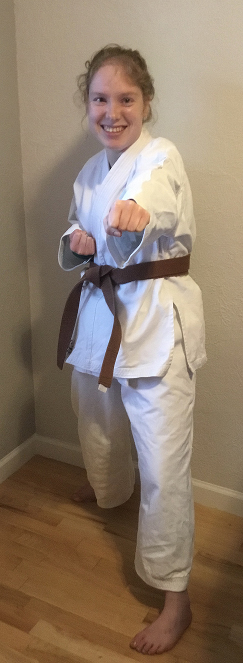 A photo of Melissa in her brown belt karate uniform. She is posing in a karate punch form, with hardwood floors and beige wall behind her.