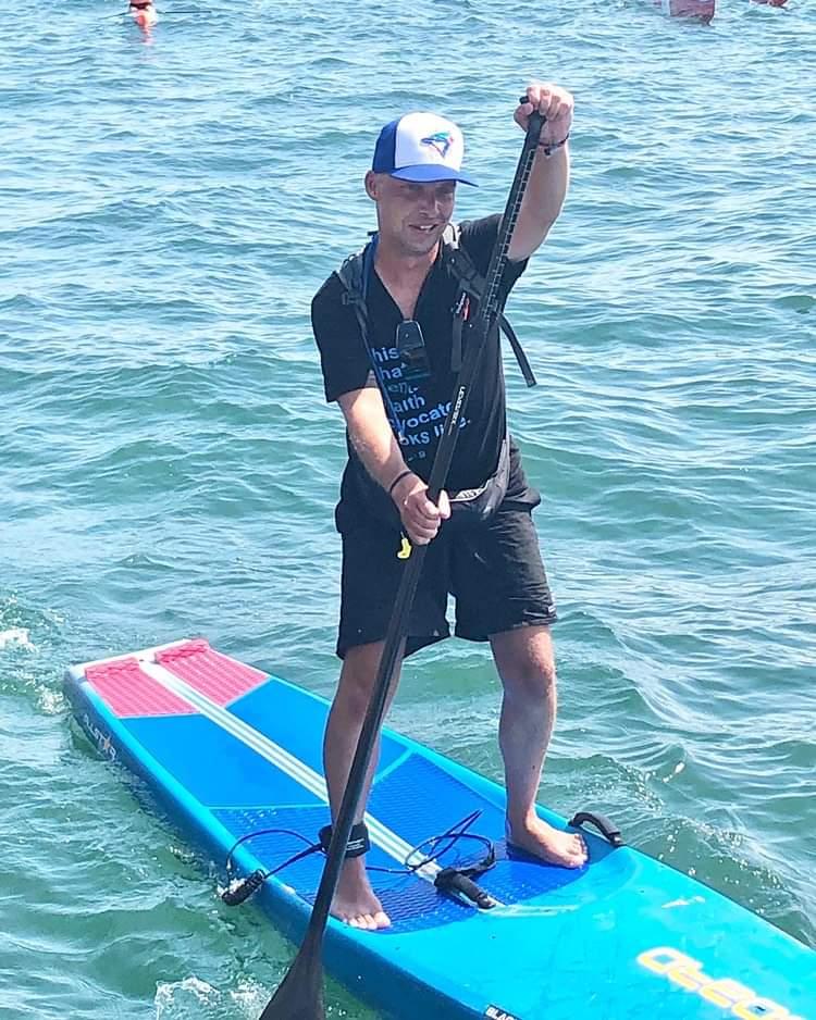 Close up shot of Mike Shoreman smiling and paddleboarding. He is wearing a black t-shirt and shorts and a Toronto Blue Jays ball cap. 