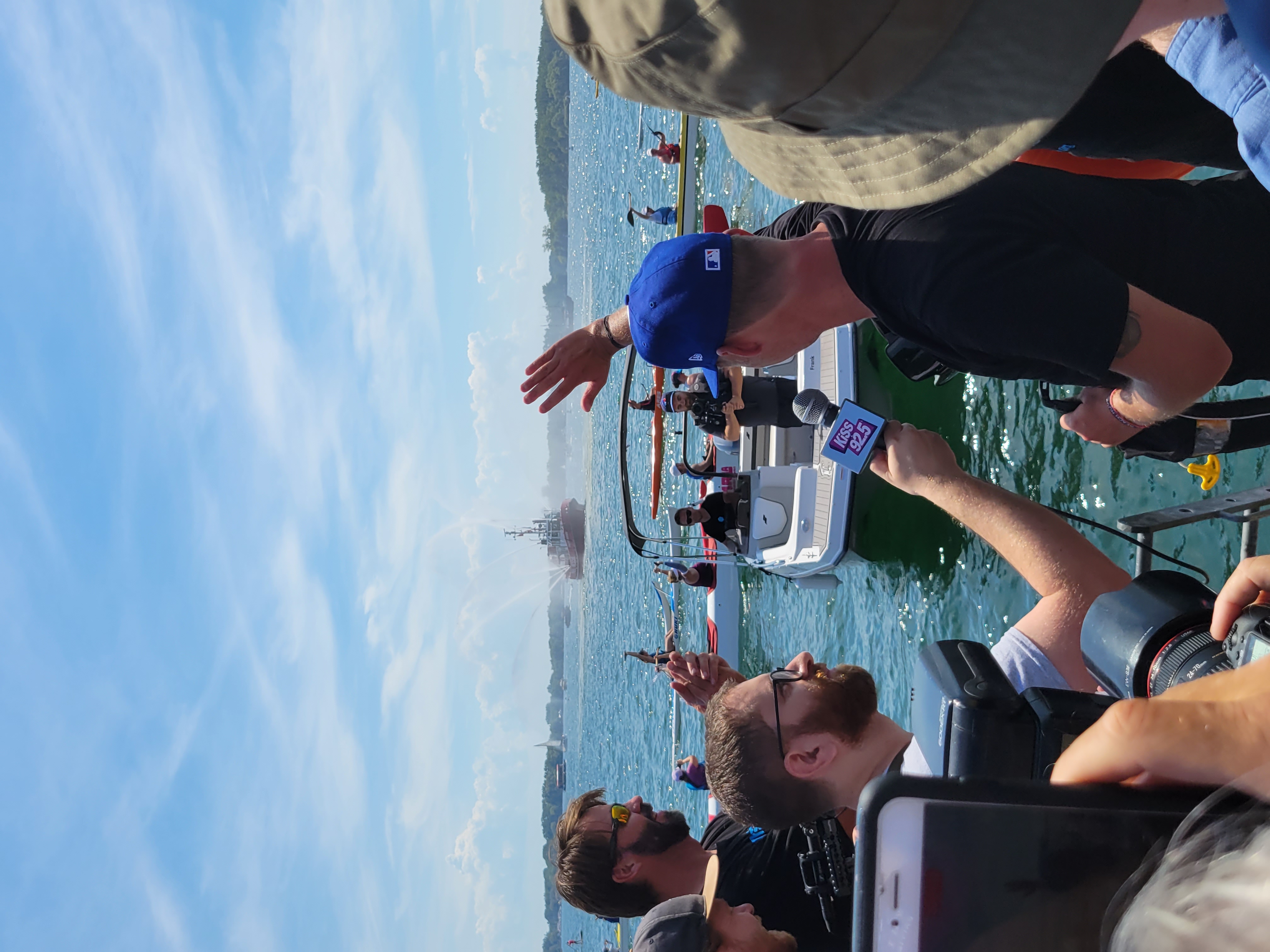 Back of Mike Shoreman's head as he is getting interviewed by a person holding a microphone. There is a crowd around Mike. He is waving to many boats on the water in front of him.