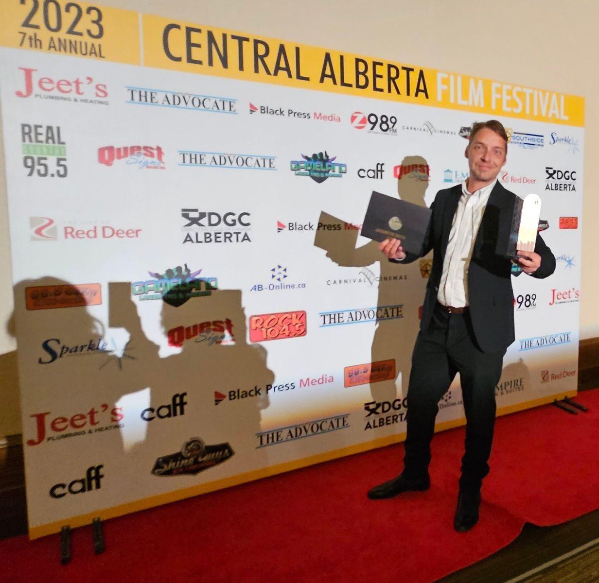 Mike Shoreman standing in front of a Central Alberta Film Festival sign holding an award and a large black envelope.