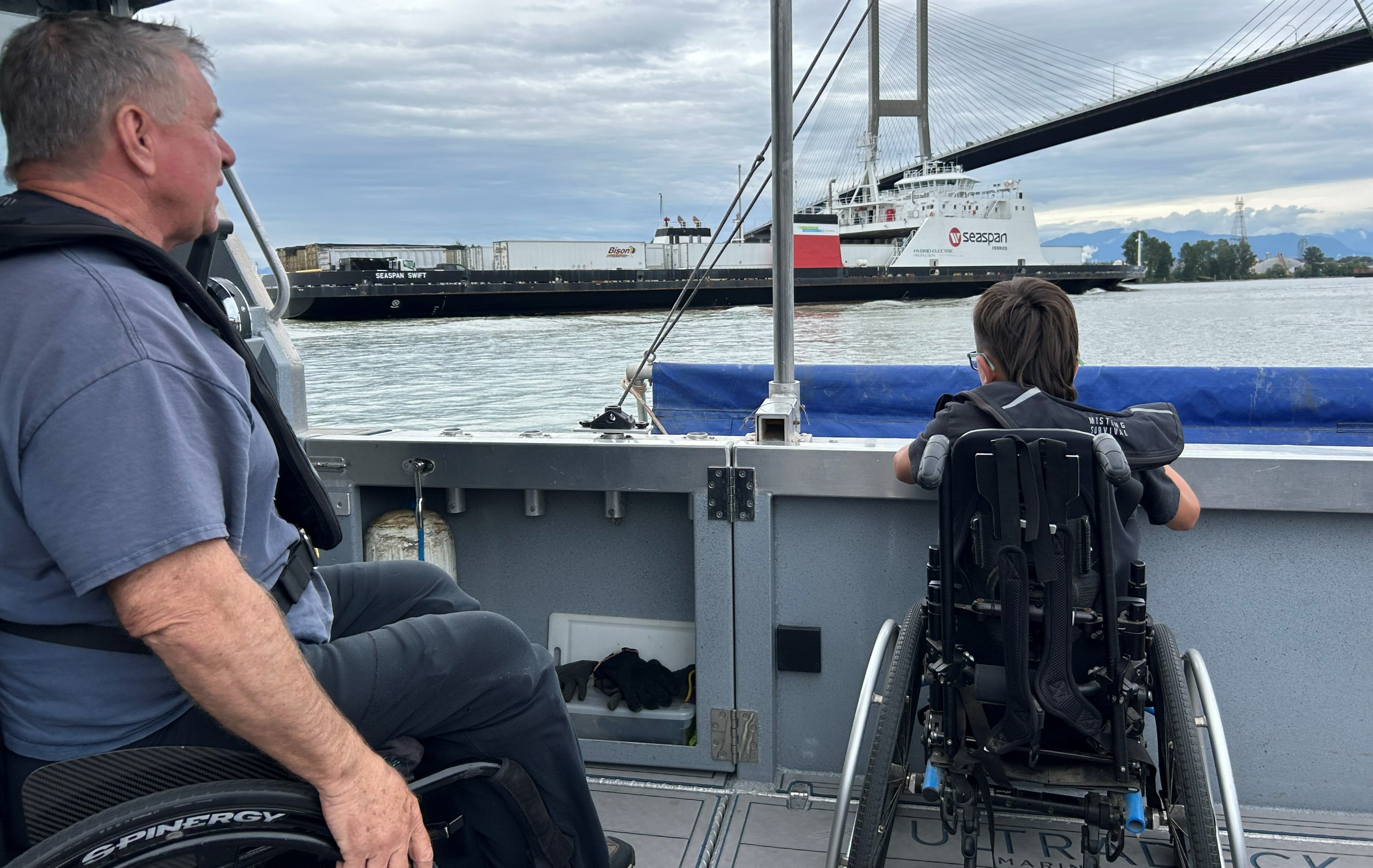 Oscar and Rick pose with four other people aboard an accessible fishing boat docked at a marina. Rick and Oscar are positioned near the centre of the group.