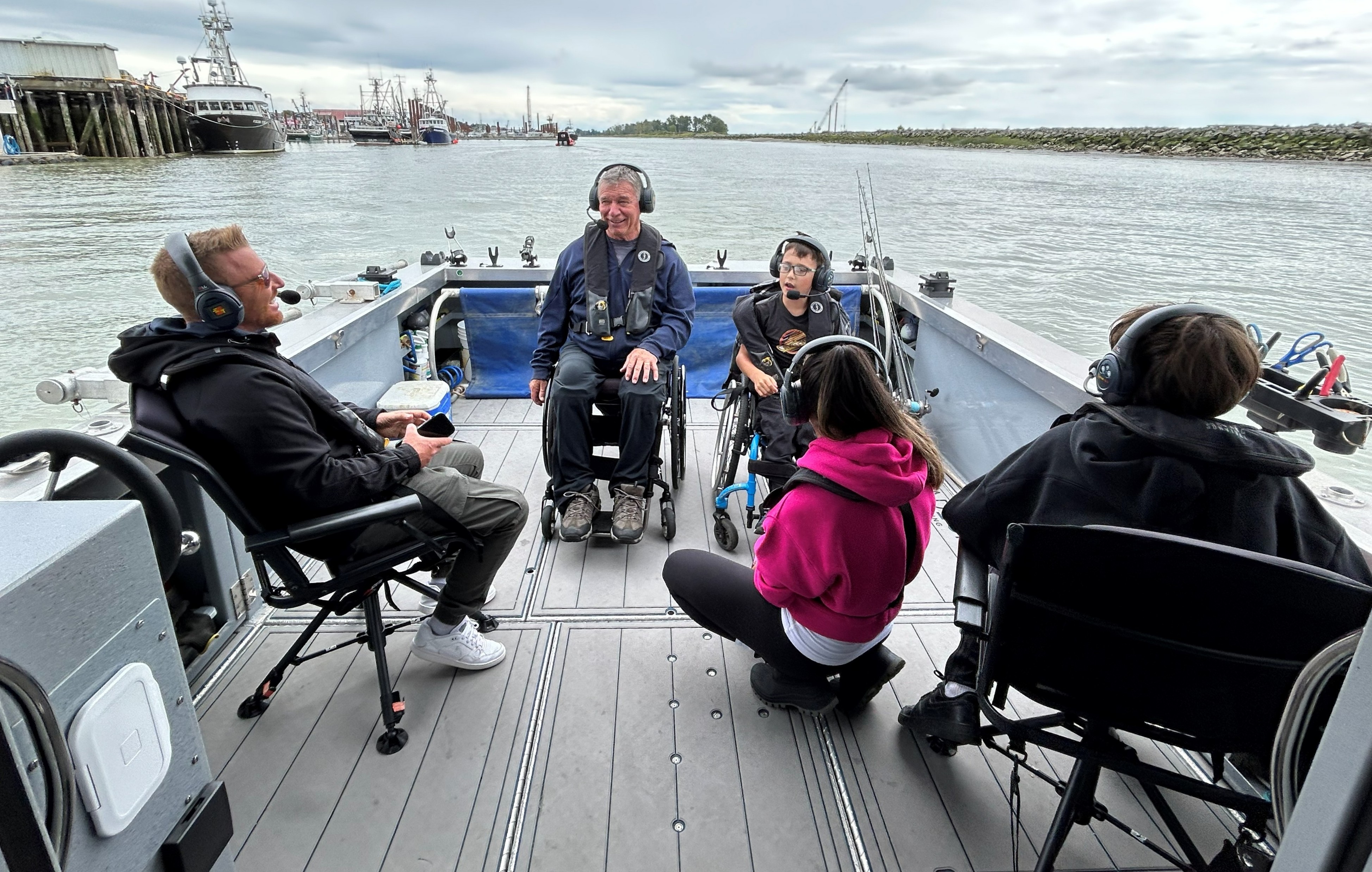 Rick and Oscar, both using wheelchairs and wearing headsets and life jackets, sit with other people aboard an accessible fishing boat in a harbour.