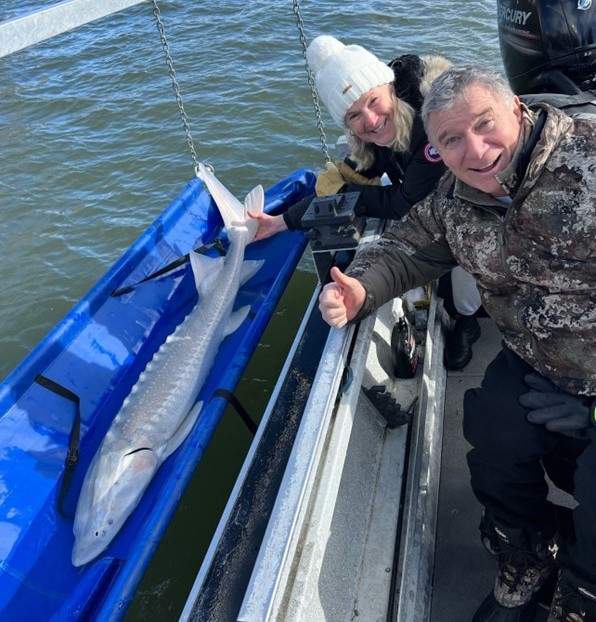 A man and a woman on a boat lean over a container holding a large fish, with water visible around them. Lorna wears a white beanie and dark jacket, and Rick Hansen wears a camouflage jacket and gloves and is using a wheelchair, giving a thumbs‑up.