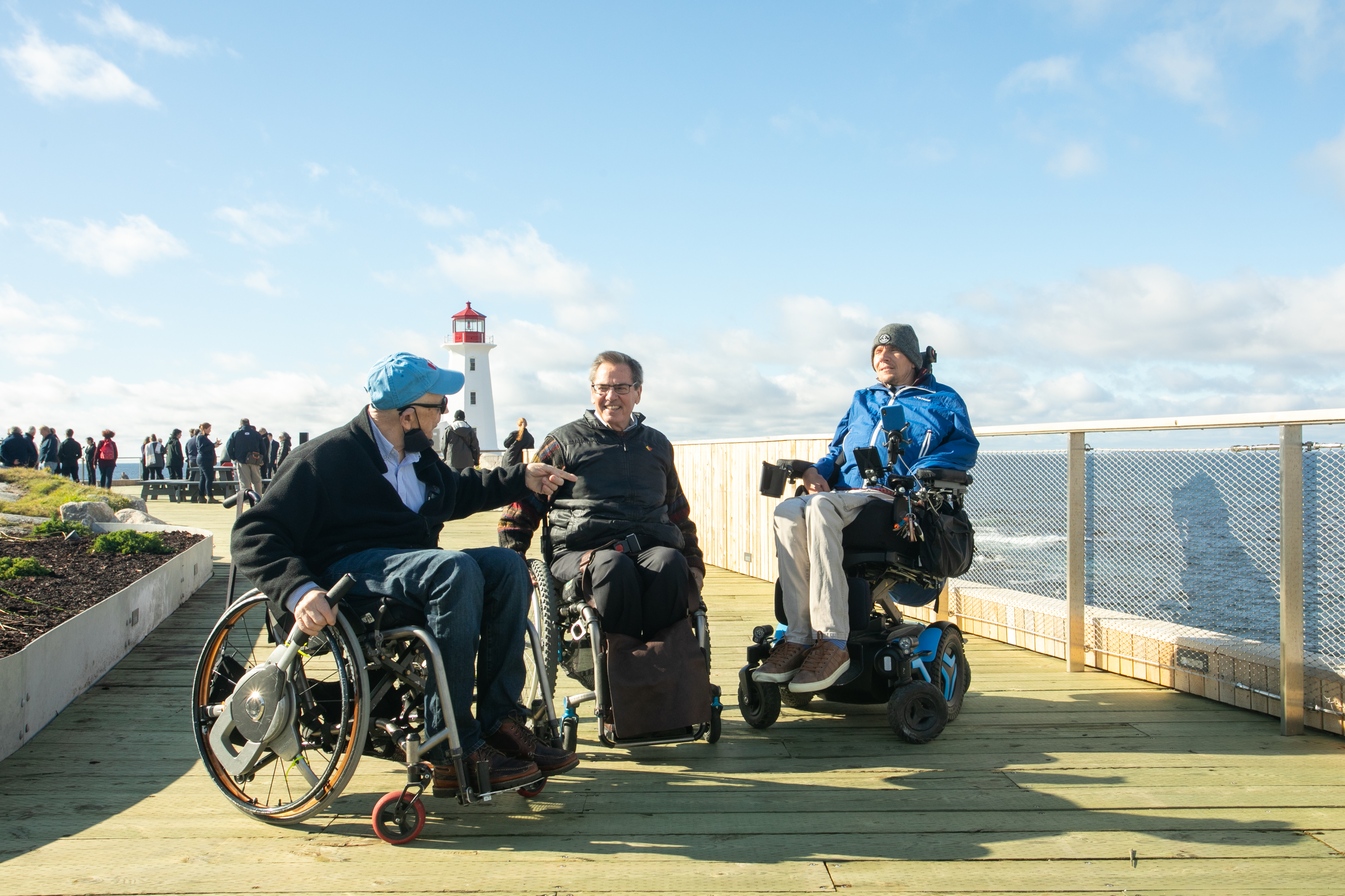 Three people who are using wheelchairs on the viewing deck at Peggy's Cove. They are facing each other and talking. Everyone is wearing jackets and long pants. The lighthouse is in the background.