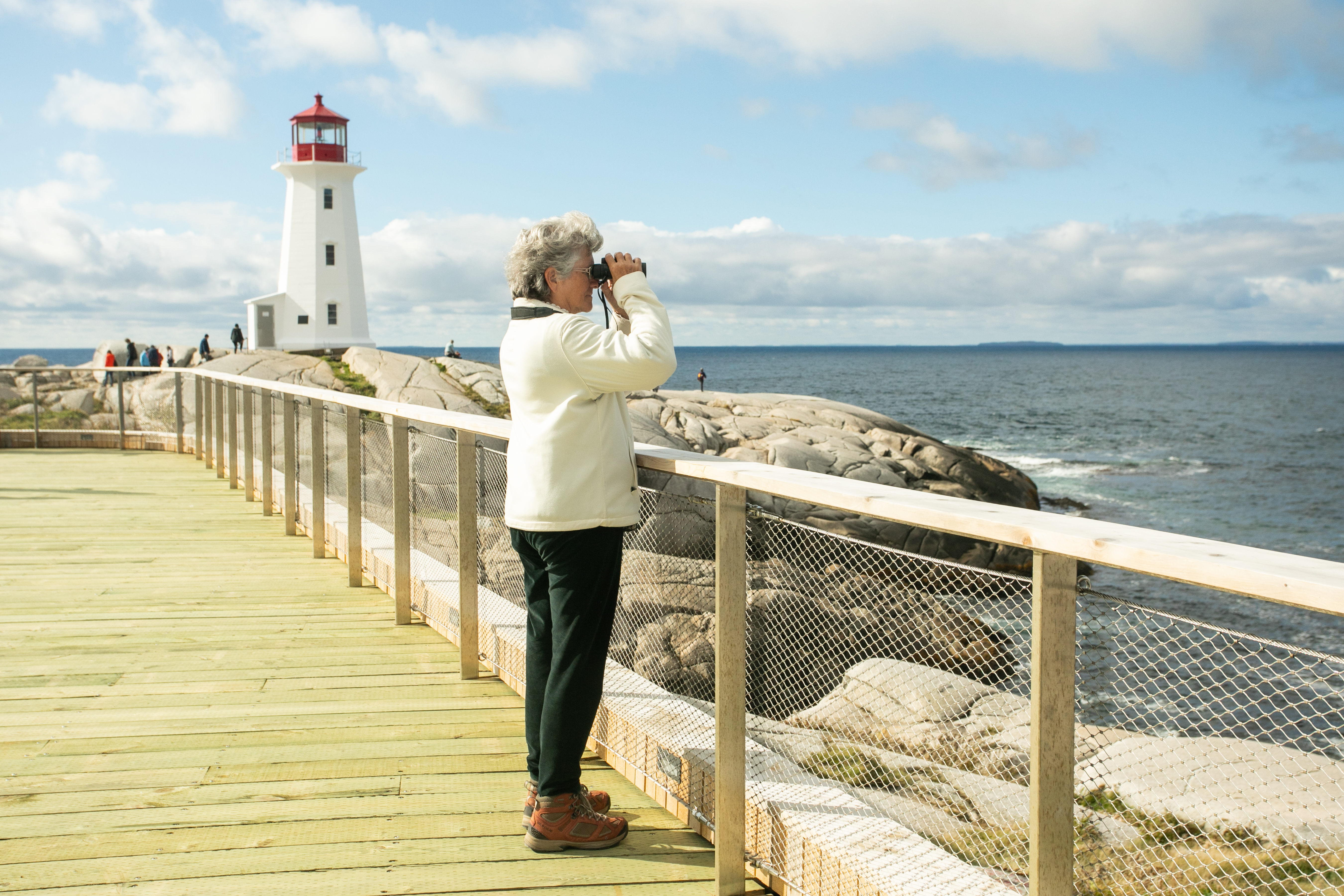 Person with short grey hair on the Peggy's Cove viewing deck using binoculars to look out at the ocean. The white and red lighthouse is in the background. 
