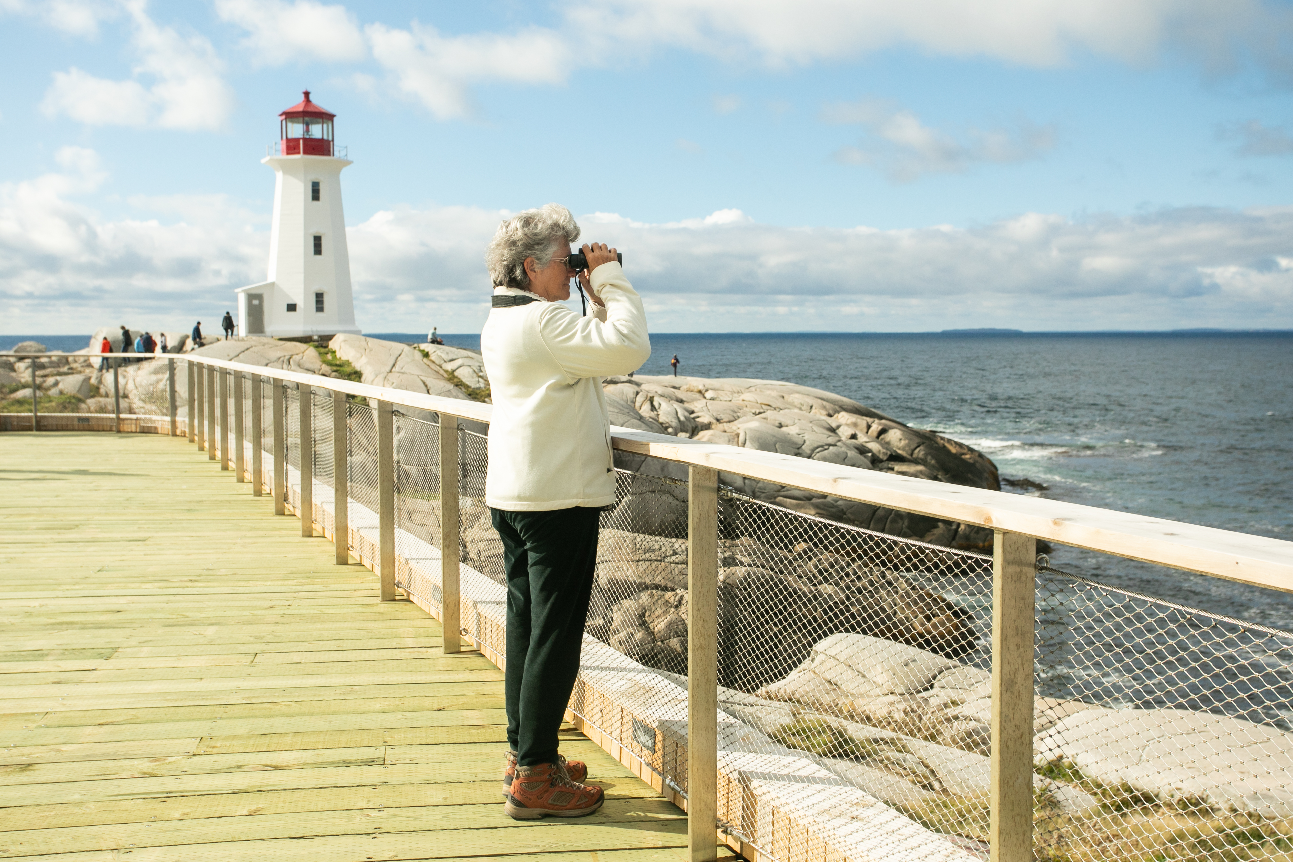 Person with short grey hair on the Peggy's Cove viewing deck using binoculars to look out at the ocean. The white and red lighthouse is in the background.